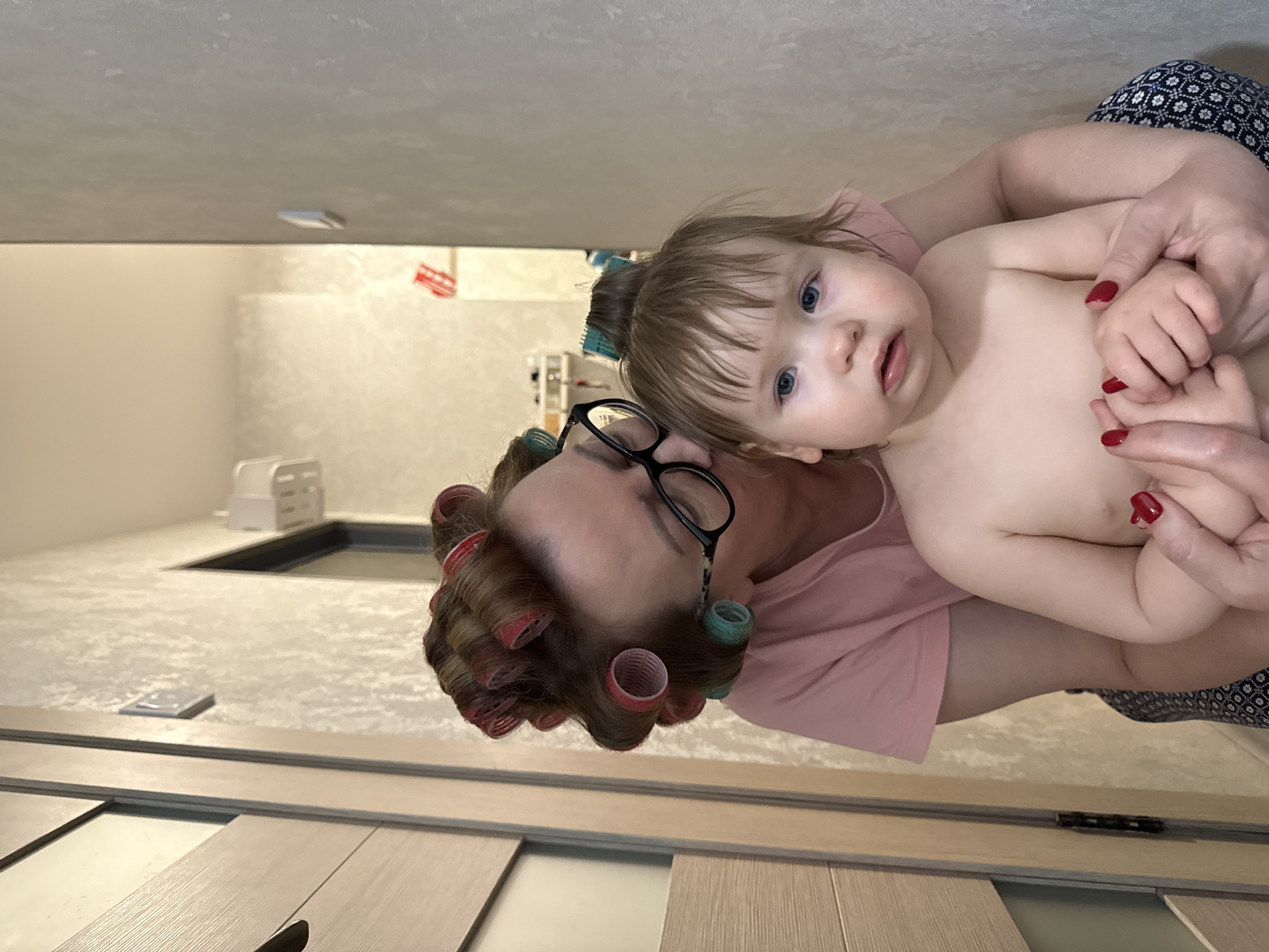 An indoor, vertical shot features a woman cradling a baby, both looking towards the viewer. The woman, with brown hair set in numerous pink and teal/green hair rollers, wears black-framed glasses and a light pink top. Her hands, adorned with red nail polish, gently hold the baby's hands. The baby, with light brown hair, striking blue eyes, and fair skin, appears shirtless and calm.
The setting appears to be a room, likely in a home in Moscow, Russia, that is under construction or renovation. Light-colored, wood-look flooring panels are visible in the foreground, some laid out, suggesting ongoing installation. In the background, there's a light-colored surface that could be a countertop, featuring a dark, rectangular cutout, possibly for a sink or appliance. A white, rectangular storage unit or small appliance is also visible further back. The walls are a light, neutral color. The lighting suggests an indoor, daytime scene. The overall atmosphere is intimate and domestic, capturing a moment between a caregiver and a child amidst a home improvement project.