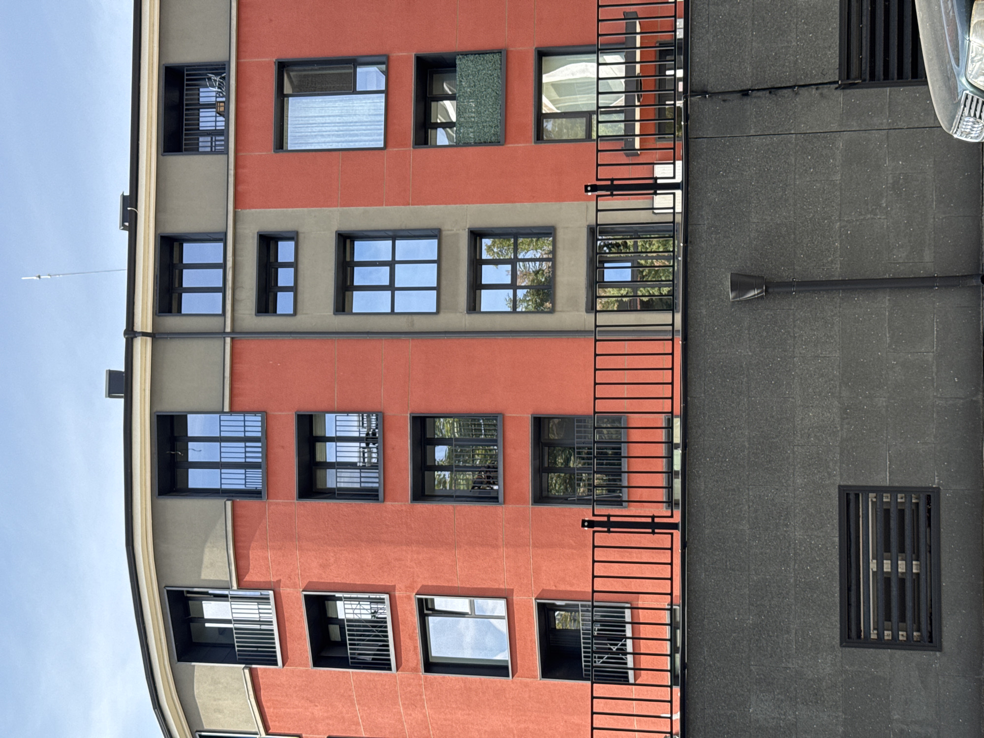 This image shows the exterior of a modern building in Plovdiv, Bulgaria. The building has a distinctive curved facade and is painted in a vibrant red and grey color scheme. Multiple floors of windows and balconies are visible, some with black metal railings and others with glass. Reflections in the windows show trees and a blue sky. In the foreground, a black metal fence runs horizontally across the image, separating the building from a dark grey concrete wall. Below the wall, there is a black vent and a downspout. A portion of a car is visible in the lower right corner. The overall scene suggests an urban residential or commercial area.
