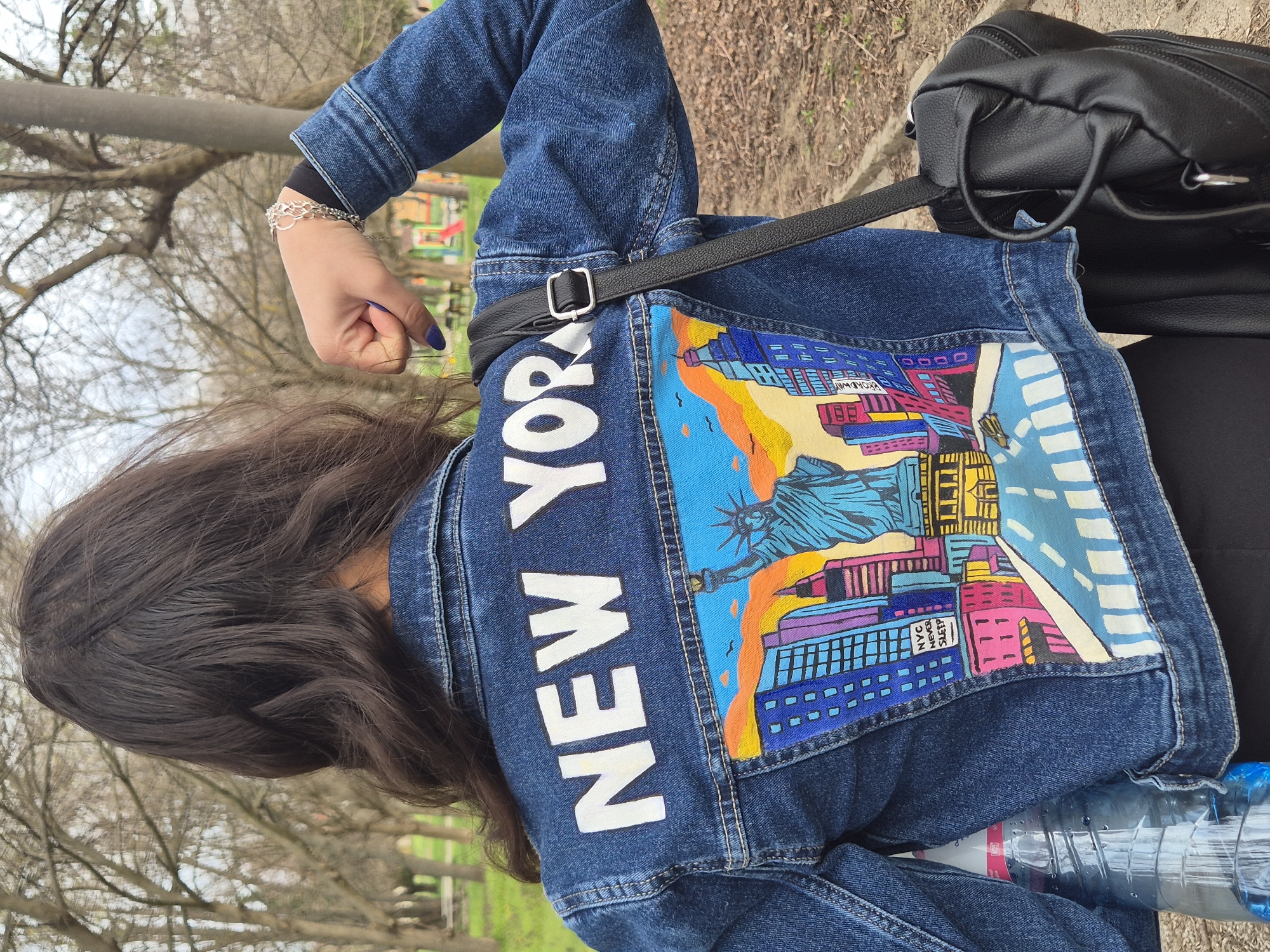 A person wearing a denim jacket with "NEW YORK" printed vertically on the left side and a colorful illustration of New York City landmarks on the back is standing in a park. The statue of liberty is visible in the center of the illustration. A black backpack hangs over the right shoulder. The person has dark hair and is wearing a bracelet on the left wrist. A water bottle is visible at the bottom of the frame. The location is in an outdoor park setting, possibly in Pavlikeni, Bulgaria.