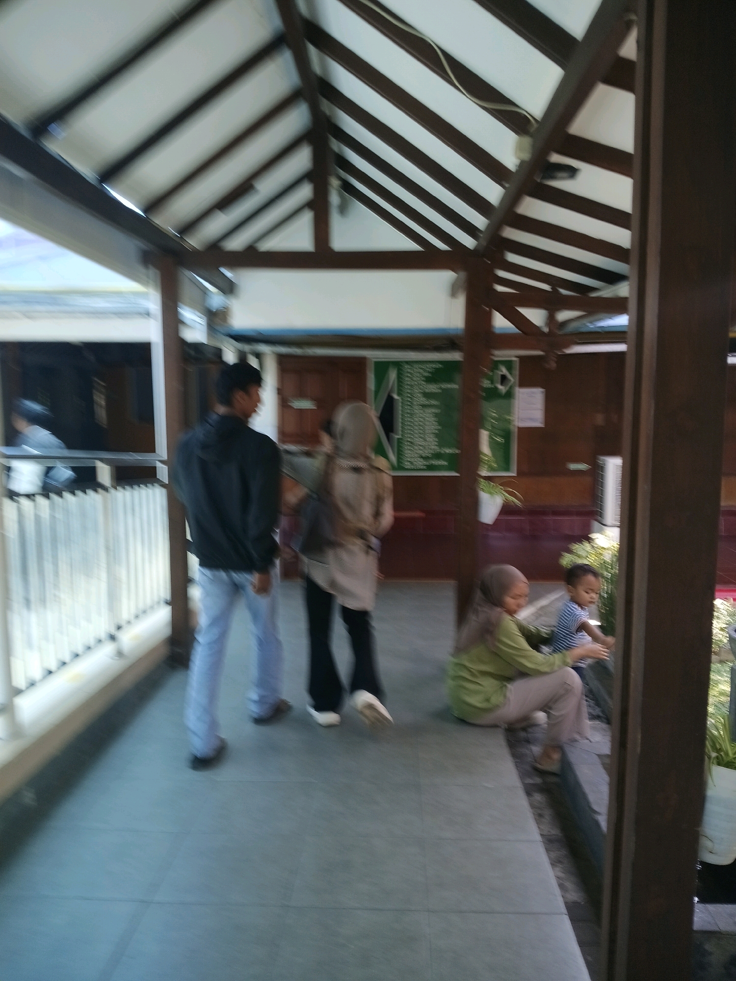 In Purwodadi, Indonesia, a blurry outdoor scene depicts a covered walkway with a dark wooden beam structure supporting a white ceiling. On the left, a man in a black jacket and light blue pants walks away from the camera. Beside him, a woman wearing a light brown hijab and a beige jacket also walks away. Further into the scene, to the right, a woman in a green long-sleeved shirt and light-colored pants sits on a low ledge, facing left. A young child in a blue and white striped shirt is seated near her. Behind them, a white air conditioning unit is mounted on a wooden wall. Green boards with white text are visible on the wall. The ground is tiled.
