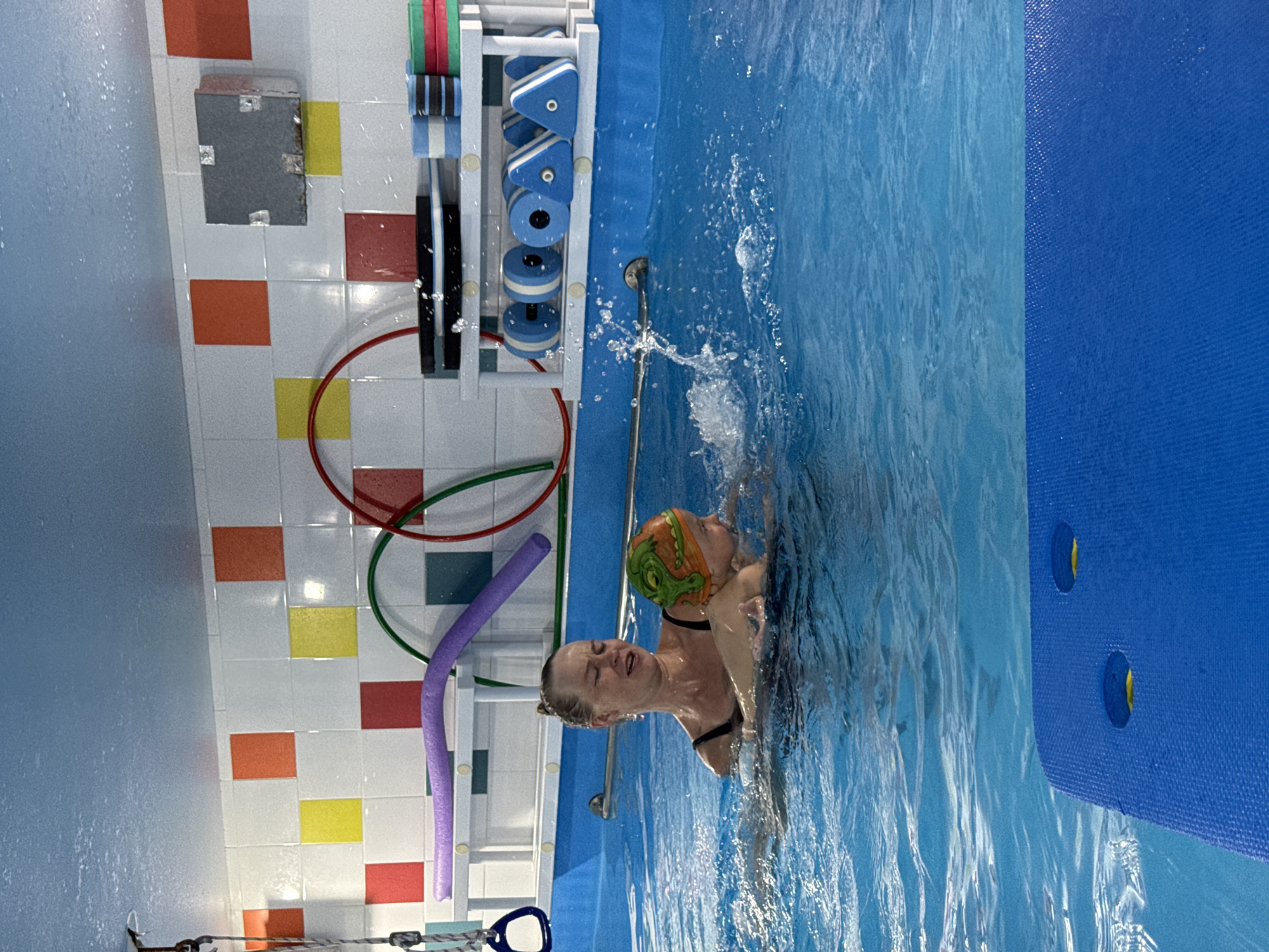 The image, set in Sochi, Russia, captures an indoor swimming pool scene featuring a woman and a child. The woman, possibly an instructor or parent, supports the young child in the water. She wears a dark swimsuit and has a focused expression, with water droplets on her face from the splashes around them. The child wears a distinctive orange and green swimming cap with an alligator or dinosaur design.
Both individuals are partially submerged in the vibrant blue pool water, which shows ripples and splashes, indicating movement. Along the tiled wall next to the pool, a white shelving unit is stocked with various swimming aids, including blue and white foam kickboards and dumbbells. A purple foam pool noodle is also visible, along with red and green hoops hanging on the wall. The wall itself is decorated with a checkered pattern of white, orange, yellow, and red square tiles, and a grey rectangular object is affixed to it. A metal handrail extends into the water from the pool's edge. The setting appears to be a facility for children's swimming lessons or water activities, illuminated by artificial indoor lighting. No visible text is present in the image.