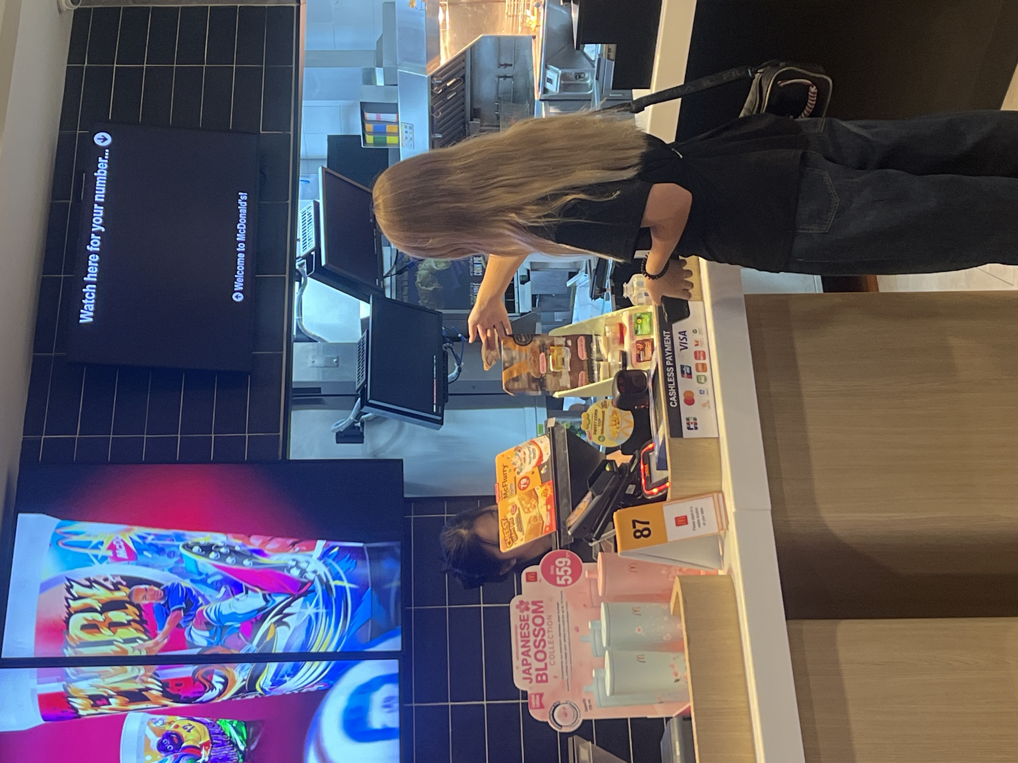 A young woman with long blonde hair is standing at a McDonald's counter, reaching out to take a food item from an employee. The employee, a young woman with dark hair pulled back, is partially visible behind the counter. Above them, a large screen displays "Watch here for your number..." and "Welcome to McDonald's!". The counter displays various McDonald's promotional items, including cups and packaging, along with a "Cashless Payment" sign featuring Visa and Mastercard logos, and a table number sign displaying "87". The environment appears to be a McDonald's restaurant, with kitchen equipment visible in the background.
