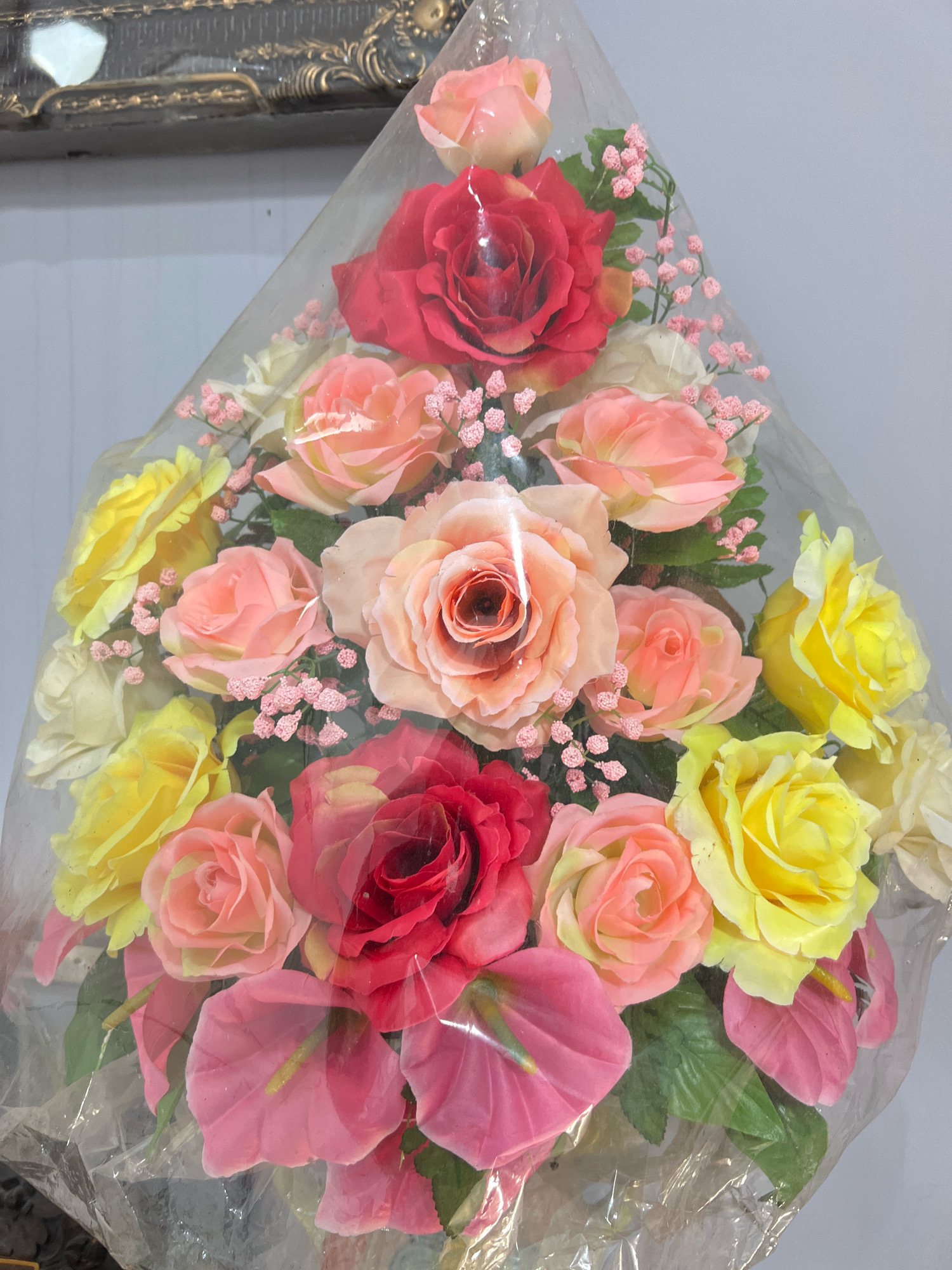 The image shows a bouquet of artificial flowers, primarily roses in shades of red, pink, and yellow, with some pink anthuriums and small pink filler flowers, all enclosed in clear plastic wrap. The bouquet is arranged in a cascading style. The background is a plain, light-colored wall and a portion of an ornate, dark-colored decorative frame. There are no people visible in the image. The scene appears to be an indoor setting, possibly a shop or home. The image quality is slightly blurry. The location in Denpasar, Indonesia, is not discernible from the visual information present in the image.