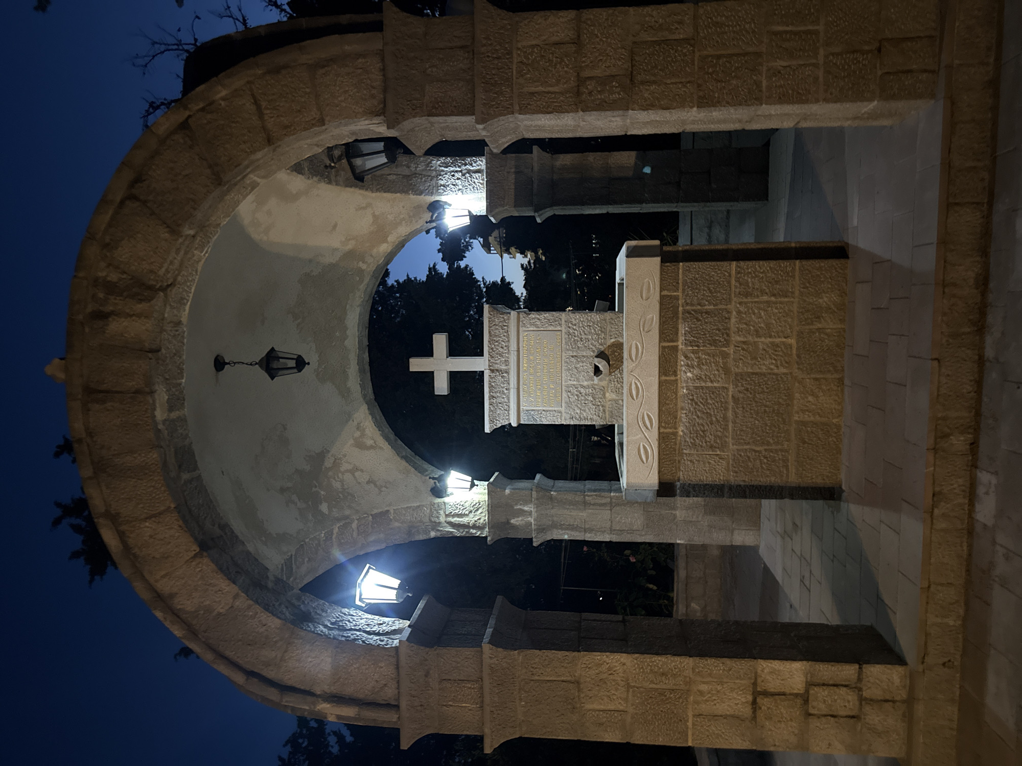 This is a night-time, eye-level, full shot of an outdoor stone structure with an arched entrance. The structure is made of rough-hewn, light brown stones and appears to be a small, open pavilion or shrine. Inside the pavilion, at the center, is a white cross atop a stone altar or monument with engraved text. A small spout is visible on the front of the monument, suggesting it may be a fountain. The ground is paved with square stone tiles. Several outdoor lanterns are mounted on the structure, providing artificial light. The background shows dark trees and a twilight sky. The scene is located in the city of Boreti, Montenegro.