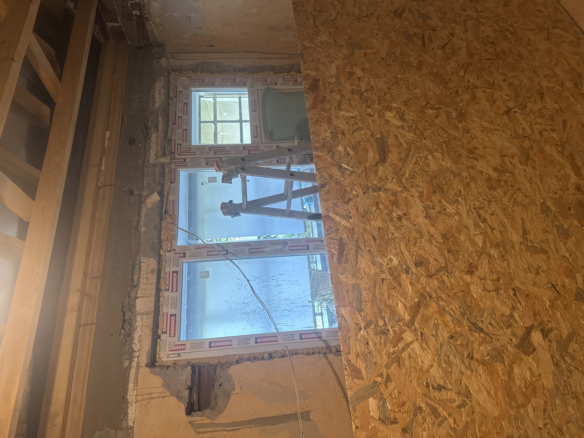 The image depicts an interior space undergoing renovation in Sarnevo, Bulgaria. The foreground is dominated by a large sheet of Oriented Strand Board (OSB) covering the floor. Above this, three newly installed, white-framed UPVC windows are visible. The window frames are sealed with tape displaying the brand "HAMMER." The two left windows are larger, while the rightmost window is narrower and taller. Through the windows, an exterior scene is vaguely discernible, including some greenery and an obscured structure. Two metallic stepladders are positioned in the midground, one in front of the middle window and another near the right window. The interior walls surrounding the windows are in a state of undress, revealing exposed brickwork, chipped plaster, and raw concrete surfaces. A single electrical wire is visible running horizontally along the left wall. Overhead, unfinished wooden ceiling joists are exposed, with some wiring or conduit running alongside them. The scene is illuminated by natural light filtering through the windows, casting some reflections and highlighting moisture or dirt on the glass panes. No individuals are present.