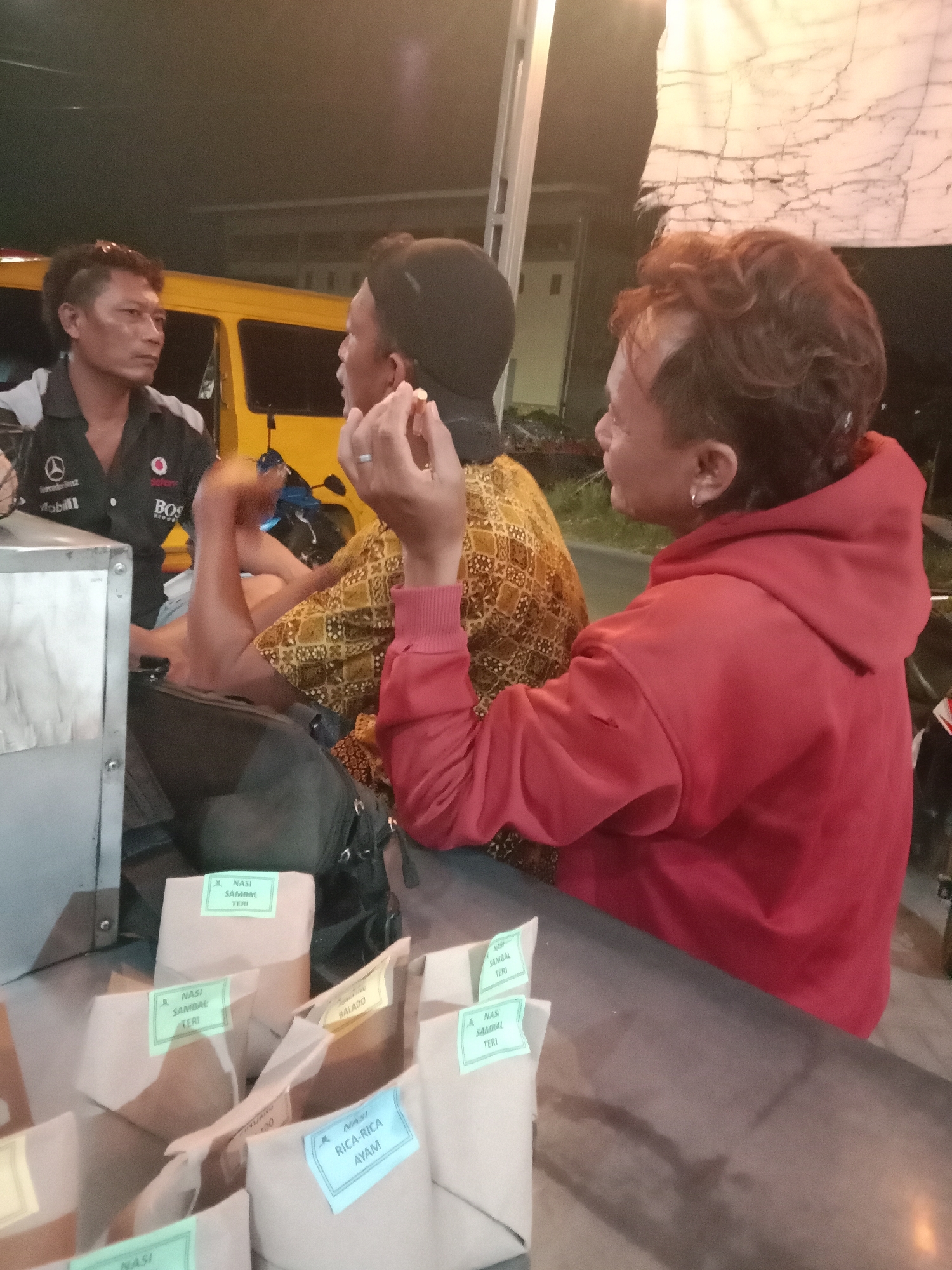 In the city of Cangkring, Indonesia, three individuals are engaged in a conversation around a food stall. The person on the left, wearing a dark shirt with logos, is looking towards the center. In the middle, a person in a patterned shirt is holding a cigarette and speaking. To the right, another individual in a red hoodie is also looking towards the center and appears to be participating in the discussion. In front of them, several food packages are neatly arranged, each with a label indicating its contents, such as "Nasi Sambal Teri" and "Nasi Rica-Rica Ayam." The background suggests an outdoor setting at night, with a yellow vehicle and a white tarp visible. The overall scene depicts a casual interaction at a local eatery.