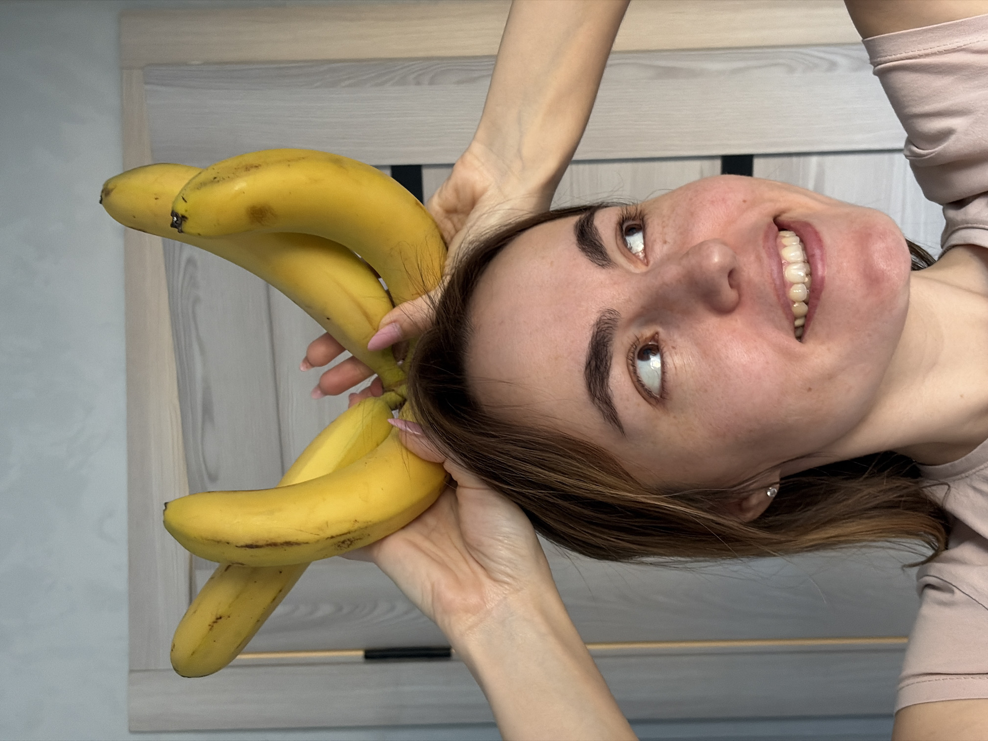 A woman holds a bunch of bananas next to her head, smiling and looking upwards. The bananas are yellow with some brown spots. She is wearing a light pink shirt and has brown hair. The background is a light grey wall and a light wood-grain door. The image is taken indoors. The location in Sarapul, Russia, is not discernible from the image.