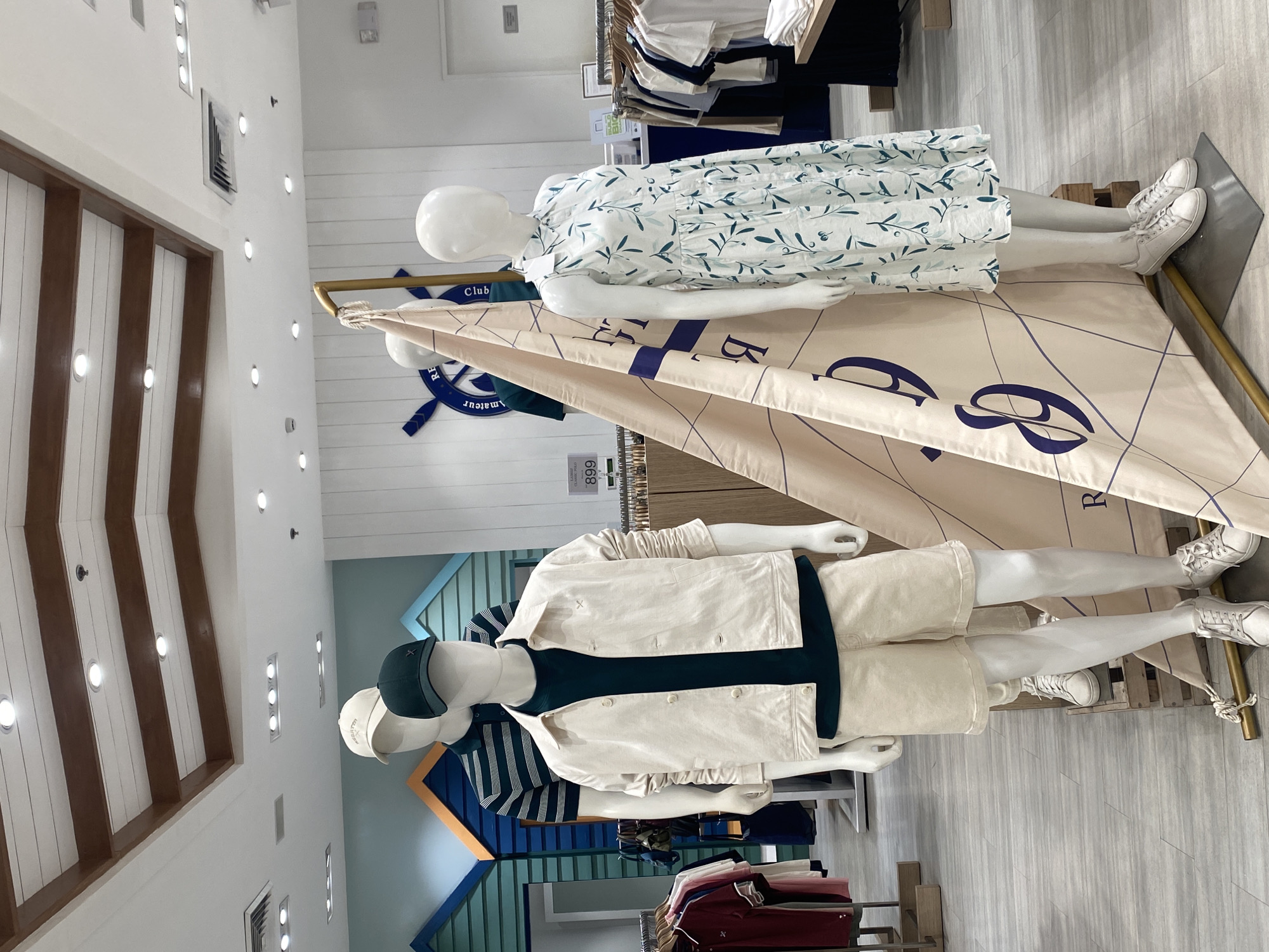 This image captures a brightly lit retail clothing store interior, featuring two mannequins displaying summer or casual wear. The setting has a distinct nautical or coastal theme.
In the foreground, a male mannequin stands, dressed in a dark green t-shirt, a light cream-colored jacket with an "X" stitching detail, and matching light cream shorts. He wears white sneakers and a dark green baseball cap with a small "X" logo. Another white baseball cap is placed on a mannequin head next to him, with some illegible text visible on it.
Behind the male mannequin, and partially obscured, a female mannequin lies reclining, dressed in a white sleeveless dress adorned with a green leafy pattern. She also wears white sneakers.
The display incorporates a large, light beige fabric sail as a backdrop, featuring large dark blue lettering that includes "Club" and partially visible "RE", possibly part of a brand name. Behind the sail, mounted on a white plank wall, is a dark blue ship's wheel logo with the word "amateur" visible below it, suggesting "Club Amateur".
The store's decor includes white plank walls, some featuring blue/green and orange accent panels designed to resemble beach huts. The ceiling is also made of white planks with exposed wooden beams, fitted with numerous recessed spotlights. The floor is a light-colored, possibly distressed wood or laminate.
Clothing racks with various garments are visible in the background and foreground. A small white tag on a clothing rack shows text "LADIES CLASSIC POLO" and the number "899", likely indicating a price. No human activity is depicted in the scene.