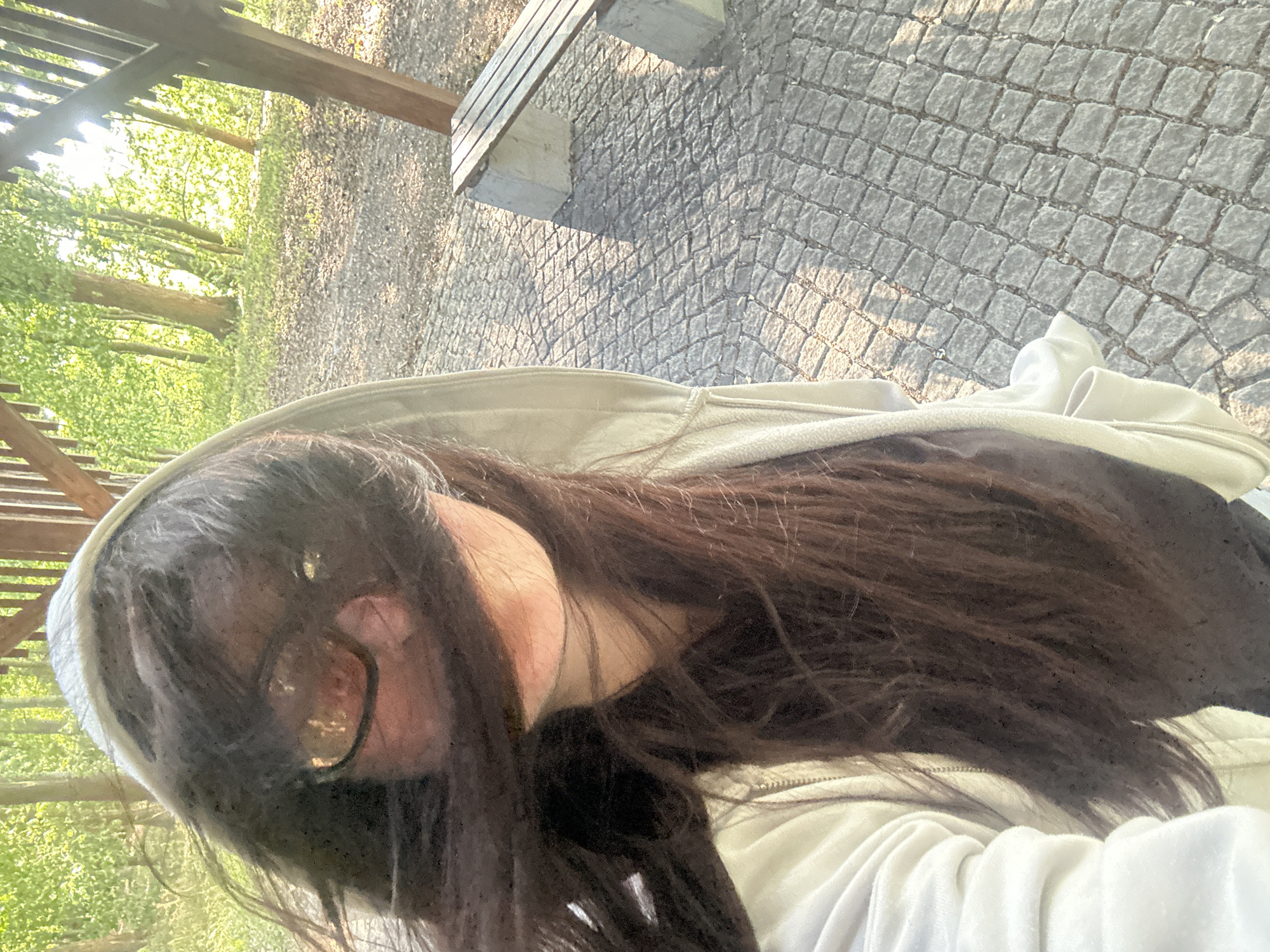 An outdoor, eye-level, selfie-style photograph shows a person wearing a light-colored hooded sweatshirt. The person has long, dark hair that is partially obscuring their face and is blown to the left. They are wearing sunglasses, and their face is turned to the right. The background consists of a wooden structure with vertical beams, a paved area with cobblestones, and trees with green foliage, suggesting a park or wooded area. In the mid-ground to the right, there is a wooden bench supported by concrete blocks. The sunlight is strong, casting shadows on the ground. The image appears to have been taken in the city of Günzburg, Germany, likely in a public park.