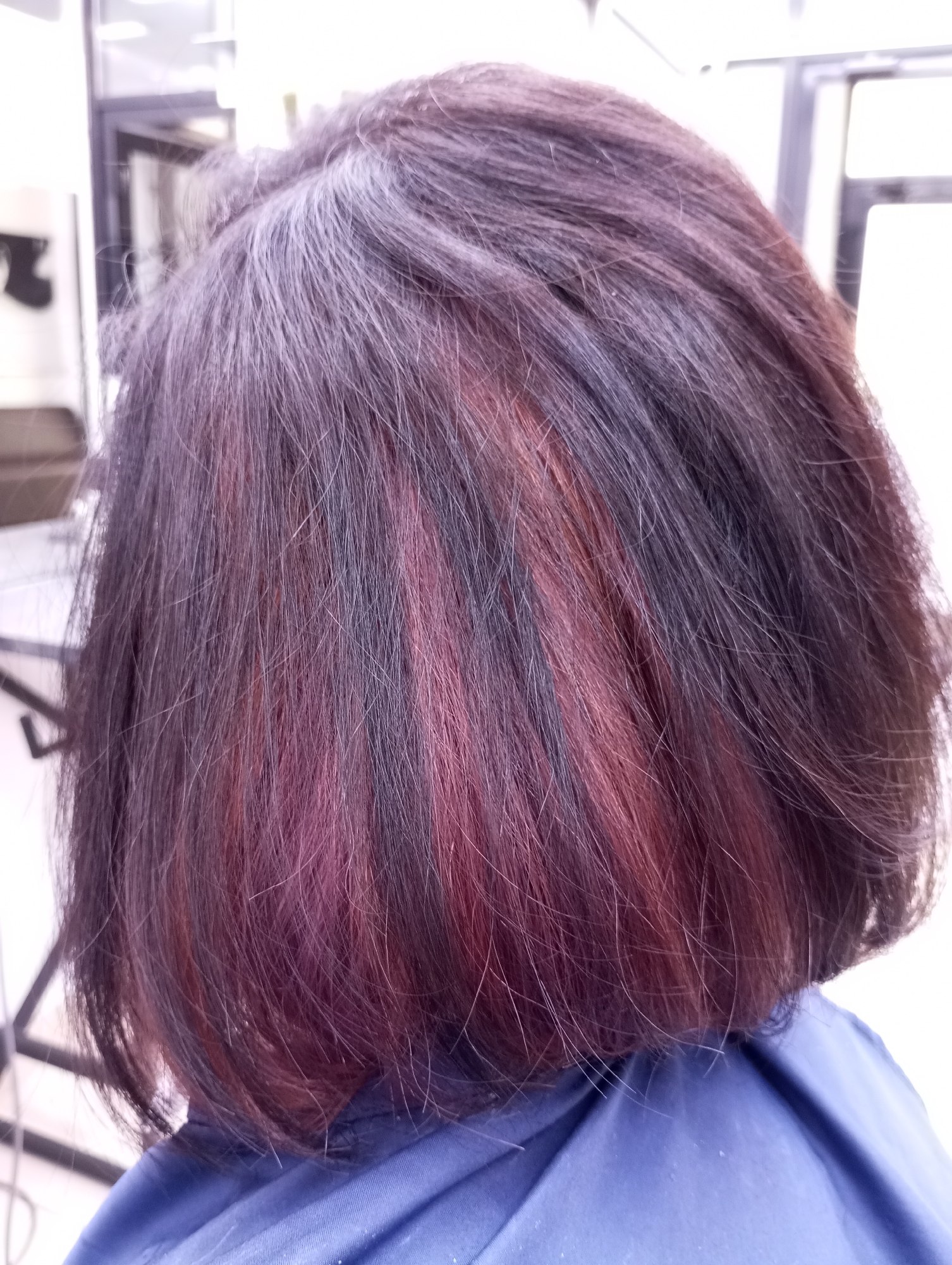 The image shows the back of a person's head with short, layered hair. The hair is primarily dark brown with reddish-purple highlights underneath. A blue garment covers the person's shoulders. The background suggests an indoor setting, possibly a hair salon, due to the presence of salon furniture and equipment. The location in Izhevsk, Russia, is not determinable from this image alone.