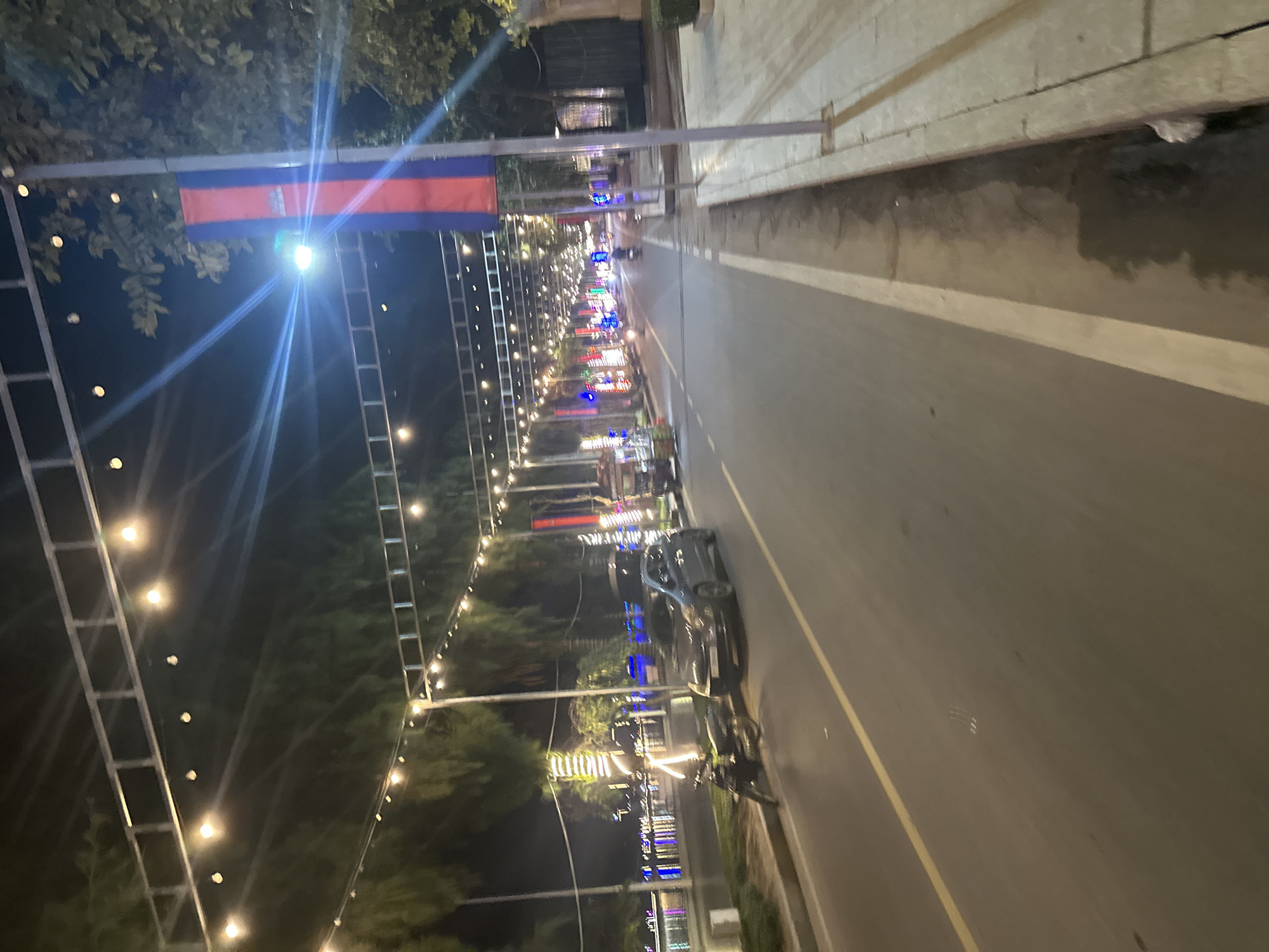 The image shows a street in Kampot, Cambodia, at night. A road runs horizontally across the frame, with a sidewalk on the right. The road is lined with trees and string lights, and a Cambodian flag is visible on a pole. A few vehicles, including a car and a motorcycle, are parked on the side of the road. The atmosphere is festive, with colorful lights illuminating the scene. There are no people clearly visible in the image.