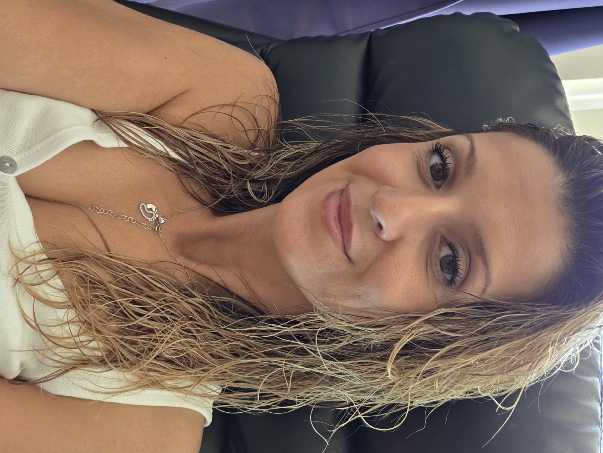 A woman with wet, blonde hair is pictured from the chest up, seated in a black chair. She is wearing a white tank top and a silver necklace with a heart and baby footprints pendant. The background is blurred, showing a hint of purple fabric and a bright window. There is no discernible indication of the location being Munich, Germany.