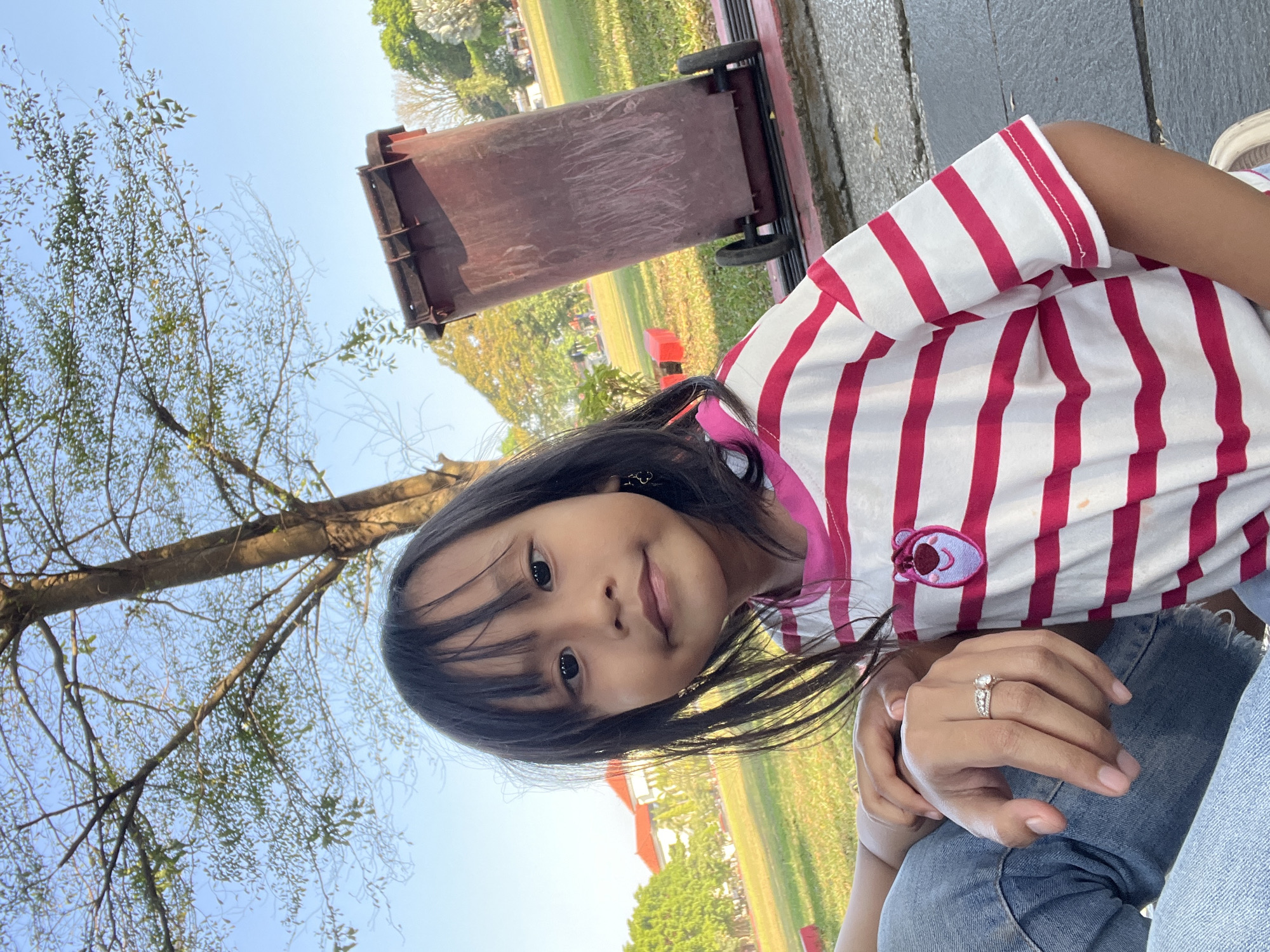 This is a vertical, close-up, outdoor selfie taken by a young girl with long, dark hair. She is wearing a white t-shirt with horizontal pink stripes and a pink trim around the collar. On the left side of her shirt, there is a patch depicting a cartoon character with pink ears and a red nose. Her right hand, adorned with a silver ring on her ring finger, is resting on her left hand. She has a playful expression with her mouth slightly open and her eyes looking directly at the camera.
The background appears to be a park or outdoor recreational area. To the left of the girl, a portion of a large tree with sparse foliage is visible against a clear blue sky. Behind her, a reddish-brown trash can with wheels is positioned next to a light-colored bench. The ground is covered with green grass, and in the distance, there are trees and buildings.
The lighting suggests it is daytime, likely morning or afternoon, with bright sunlight. The overall mood seems cheerful and relaxed, possibly indicating a casual outing or a moment of play. There are no visible text elements in the image. The context provided indicates the location as Purwodadi, Indonesia.