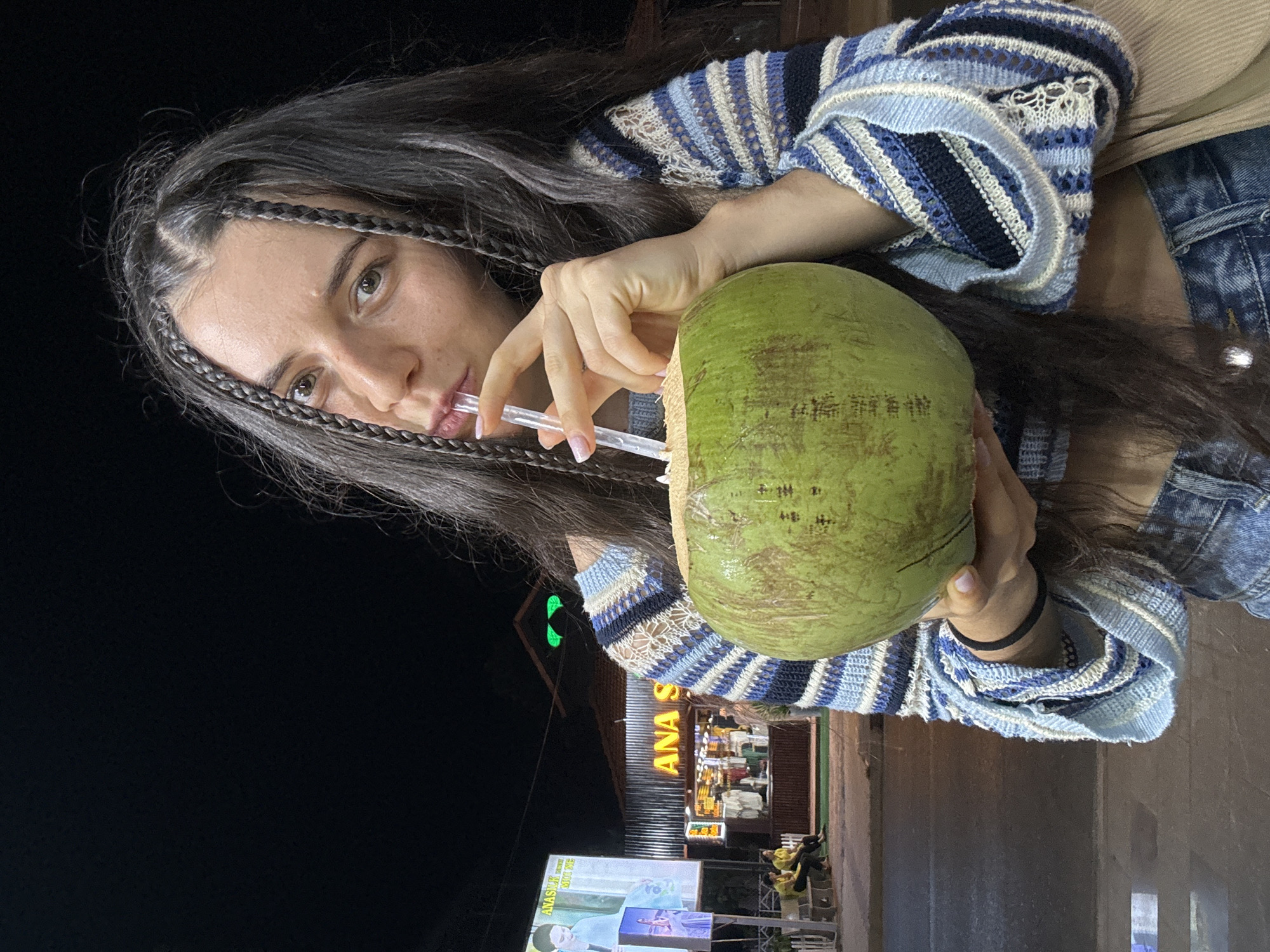 This media file captures a young woman enjoying a fresh coconut at night in Mũi Né, Vietnam. The image is taken from a slightly low perspective, looking up at the subject, with a dark night sky in the upper portion of the frame.
The central subject is a woman with long, dark hair, styled with several thin braids framing her face. She has light-colored eyes and looks directly towards the viewer with a neutral expression. She is wearing a distinctive blue, white, and dark blue striped knit top, possibly a cardigan, featuring a textured or crochet pattern. A lighter garment is visible underneath. Hints of denim clothing are seen on her lower right side, and a black band or bracelet is on her right wrist. Her fingernails are manicured with a light polish.
She is holding a large, unhusked green coconut with both hands, positioned near her face. A clear plastic straw is inserted into the coconut, and she is actively drinking from it.
The background reveals a street scene illuminated by artificial lights. A prominent sign for "ANA S" is visible on a building, suggesting a shop or eatery. Further back, a large billboard displays text, including "ANAS M.I.C...", along with an image of a man. The setting indicates an outdoor, commercial area bustling at night.
