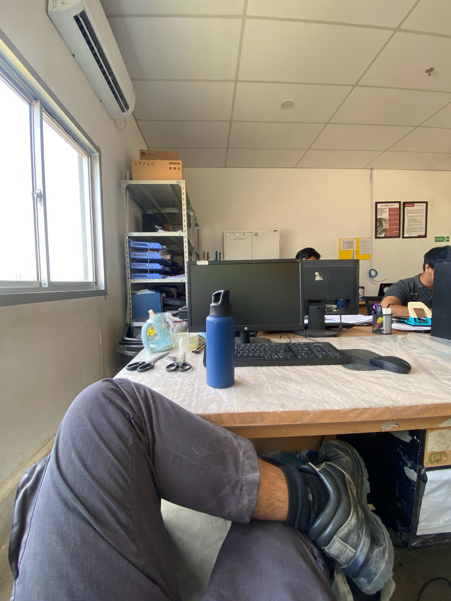 This is a medium-angle, eye-level shot taken indoors, likely in an office or workshop setting in Batam, Indonesia. The foreground is dominated by the lower half of a person's body, with their legs crossed and one foot resting on the floor. They are wearing dark grey trousers and black work boots. The person's right leg is bent at the knee, and their left leg is extended forward.
The middle ground of the image features a cluttered desk. On the desk, there is a large black computer monitor, a keyboard, a mouse, a blue water bottle, and a pair of scissors. To the left of the desk, a white shelving unit is visible, filled with various items including blue trays and boxes. A window with a grey frame is on the far left, letting in natural light.
In the background, other office desks and workers can be seen. Two individuals are partially visible, seated at their workstations. The environment appears to be a functional workspace, with fluorescent lighting on the ceiling and what appear to be informational posters on the wall. The overall impression is one of an active, albeit somewhat disorganized, work environment.