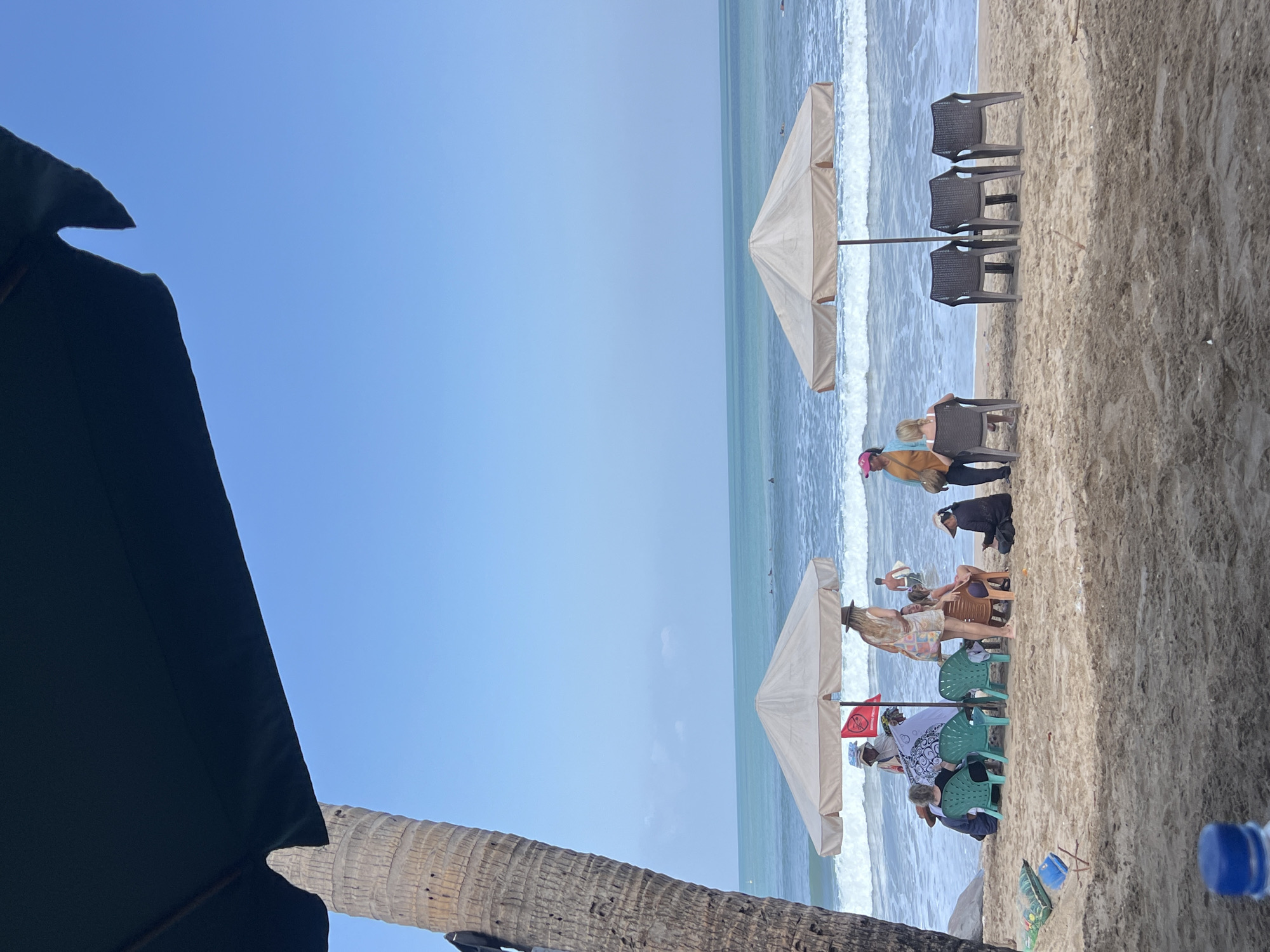 A medium shot captures a beach scene in Kuta, Indonesia, under a bright, clear blue sky. In the foreground, a large, dark umbrella partially obscures the top left of the frame, with the textured trunk of a palm tree visible to its left. The beach is sandy with some scattered debris. In the middle ground, several beach umbrellas and chairs are set up. Under one beige umbrella, a group of people are gathered, some seated on chairs and others on the sand. One person stands and holds a red flag. To the right, another beige umbrella shades three empty dark plastic chairs. The ocean is visible in the background with gentle waves breaking on the shore. Several individuals are in the water, some appearing to be surfing. The overall atmosphere is relaxed and typical of a beach resort.
