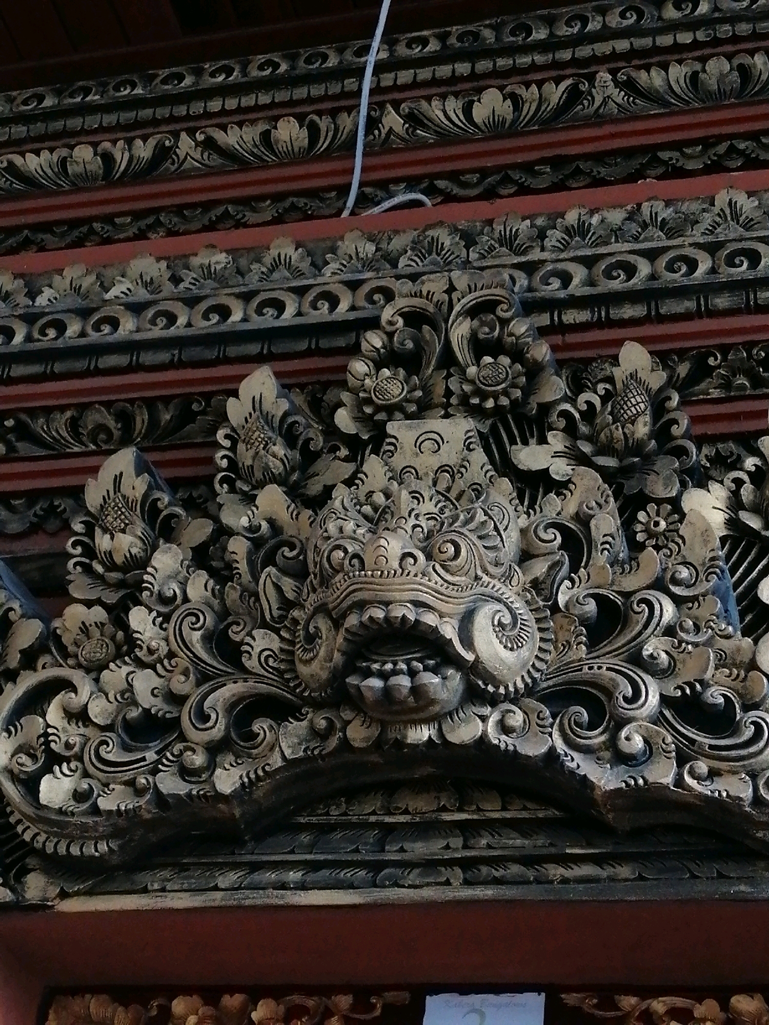 A close-up, eye-level, vertical shot captures ornate, golden-hued wood carvings adorning the exterior of a building in Ubud District, Indonesia. The carvings are intricate and detailed, featuring a central grotesque mask with bared teeth and prominent fangs, surrounded by floral motifs and swirling patterns. Bands of carved designs run horizontally across the frame, creating a textured and layered effect. The color palette is dominated by dark browns, blacks, and accents of gold, suggesting an aged and traditional aesthetic. A thin white cable is visible extending from the top of the frame downwards, a modern element juxtaposed against the ancient artistry. The background consists of deep red surfaces, possibly walls or supporting structures, and additional carved elements are visible at the bottom of the frame. The lighting is natural, highlighting the texture and depth of the carvings.