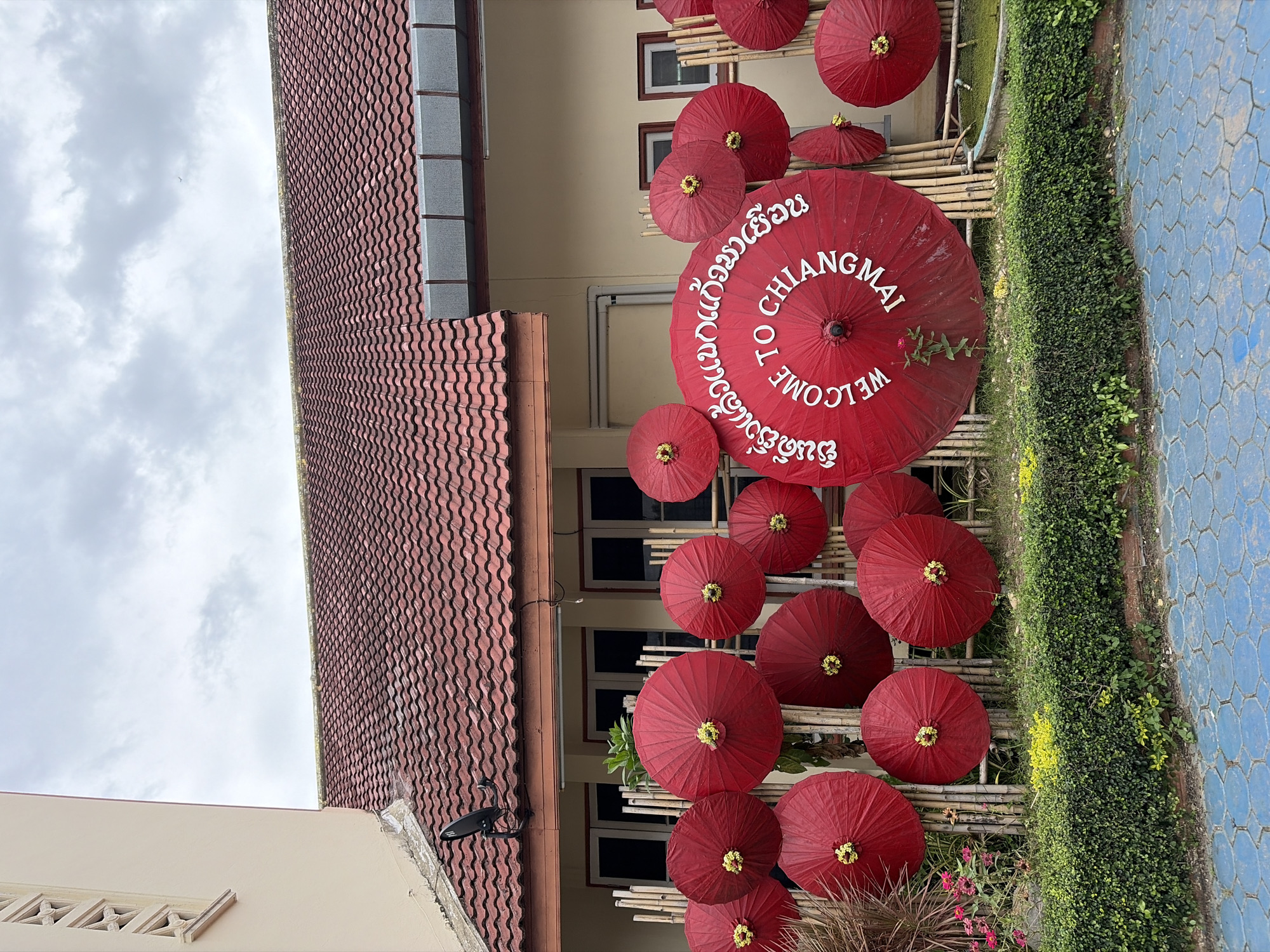 This image depicts an outdoor scene in Chiang Mai City Municipality, Thailand. The focal point is a display of red, traditional Thai paper umbrellas. One large umbrella in the center has white text written in a circular pattern, which reads "WELCOME TO CHIANGMAI" in English and what appears to be Thai script. The umbrellas are arranged in front of a building with a red tile roof and light-colored walls, with multiple windows visible. The umbrellas are supported by bamboo poles. In front of the display, there is a hedge of green leafy plants and some yellow flowers. The ground in the foreground is paved with blue hexagonal tiles. The sky above is overcast with some clouds.