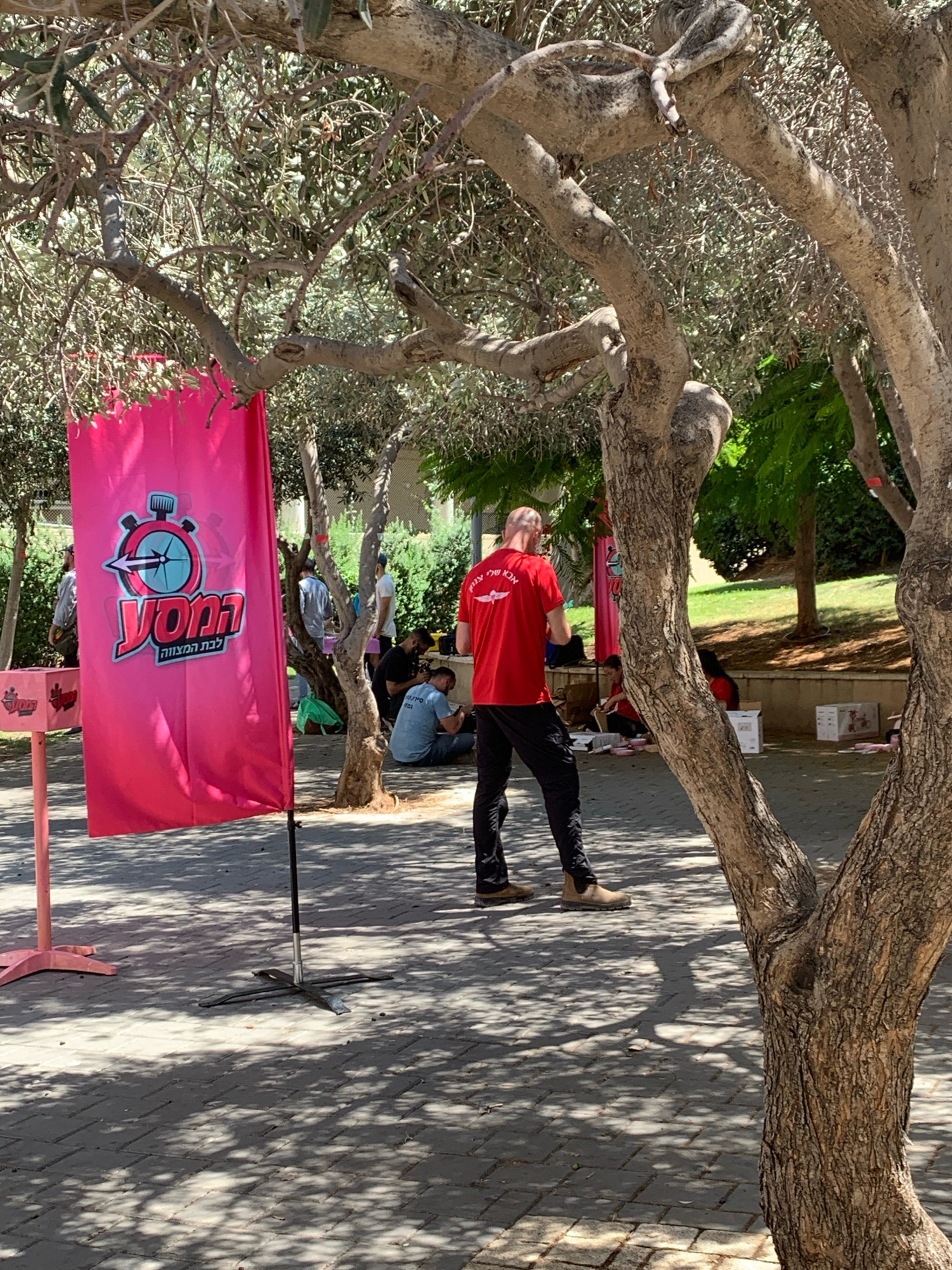 The image shows an outdoor scene, likely a park or public space, with people gathered. In the foreground, a large pink banner with a stopwatch logo and Hebrew text is prominently displayed. A man in a red shirt and black pants stands facing away from the camera, looking towards a group of people seated on the ground. Other individuals are visible in the background, some standing and others sitting, with a few boxes and bags scattered around. The area is shaded by trees, and the ground is paved with interlocking bricks. The presence of the "YONIF" (יוניף) banner suggests this might be an event organized by that entity. The Hebrew text on the banner reads "לבת המצווה" (lebat hamitzvah), meaning "for the Bat Mitzvah." This indicates a celebration for a young girl's coming-of-age ceremony. While the exact location in Tel Aviv isn't specified, the scene depicts a casual outdoor gathering, possibly in a community park or an event space within the city.