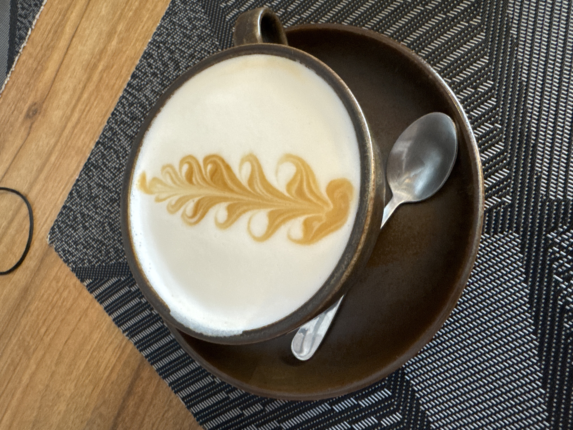 A high-angle, medium shot features a cup of latte art on a brown saucer with a silver spoon, placed on a placemat with a dark gray and white geometric pattern. The saucer and cup have a rustic, dark brown glaze with a slightly textured surface. The latte art depicts a feathery or leaf-like design in a darker brown color, contrasting against the white milk foam. The handle of the cup is visible to the upper left. The silver spoon rests on the saucer to the right of the cup. Part of a light brown, wooden surface is visible on the left side of the frame. The scene is indoors with likely artificial lighting, given the clarity and even distribution of light. It is not possible to determine the location in Tashkent, Uzbekistan, from this single image.