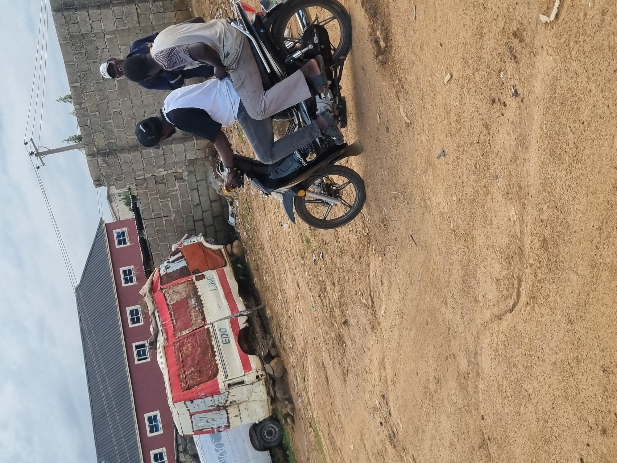 In Jos, Nigeria, an outdoor scene depicts a dusty, unpaved area with debris scattered across the ground. A red and white van, appearing derelict, is propped up on rocks. A motorcycle with two occupants, one driving and the other as a passenger, is positioned in the foreground, facing right. The driver is wearing a black cap, a white t-shirt, and grey pants. The passenger is wearing a patterned t-shirt and grey pants. In the background, a brick wall separates the scene from a red building with multiple windows and a grey roof. A utility pole with wires is also visible. Three men are standing behind the brick wall, one wearing a white cap and smiling, while the other two are partially obscured. The sky is overcast.