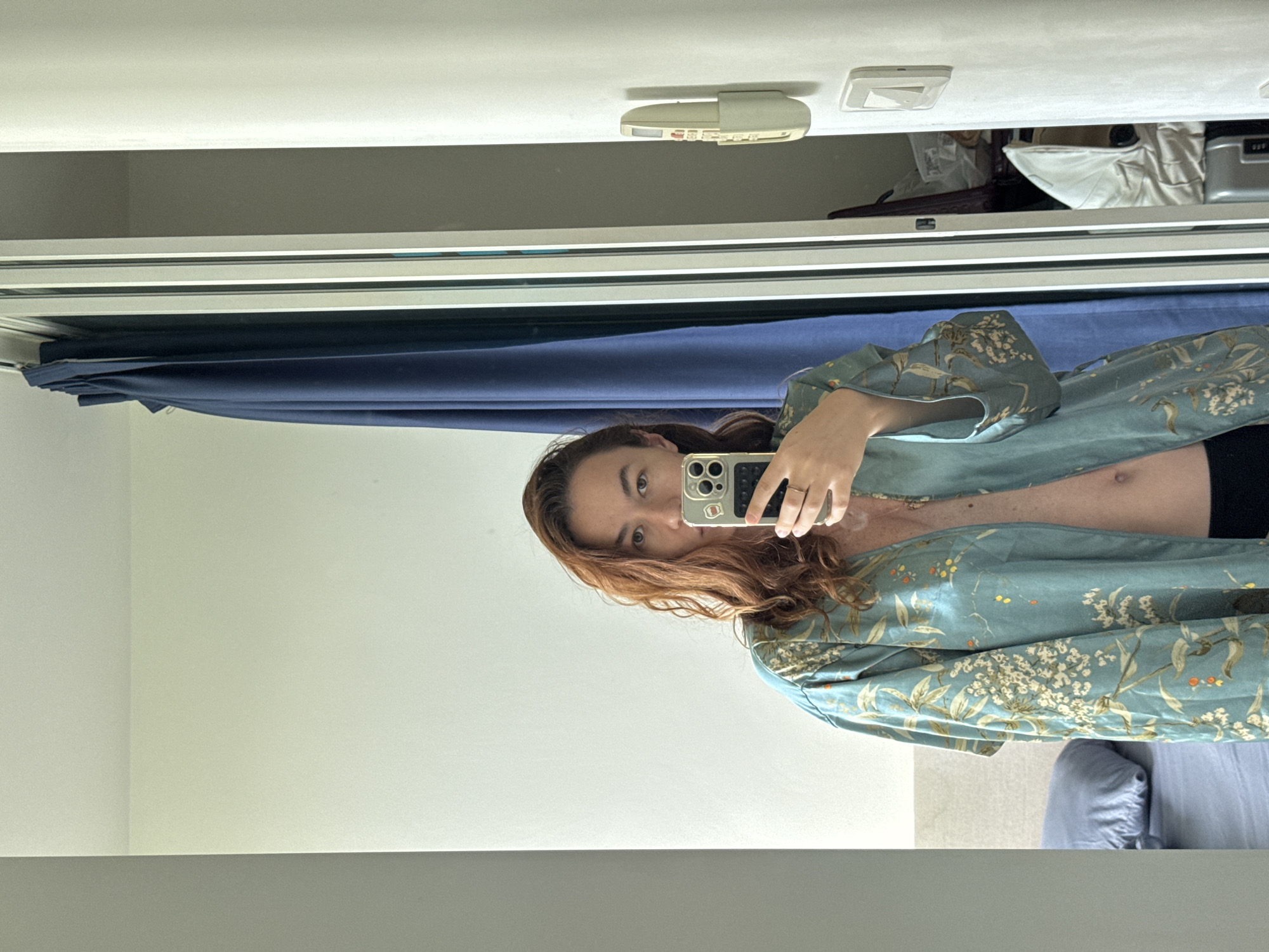 The image shows a full-body mirror selfie taken by a woman indoors. She has long, wavy auburn hair and is wearing a light blue kimono-style robe with a floral and bird pattern. The robe is open, revealing her chest and midriff. She is holding a smartphone in her right hand, pointed at the mirror. The background shows a white wall, a blue curtain, a closet with items inside, a bedside table with a pillow, and a part of a bed. There is a light switch and a remote control mounted on the wall to the right. The setting appears to be a bedroom in an apartment or hotel. There is no visible indication of the location being in Ratsada, Thailand.