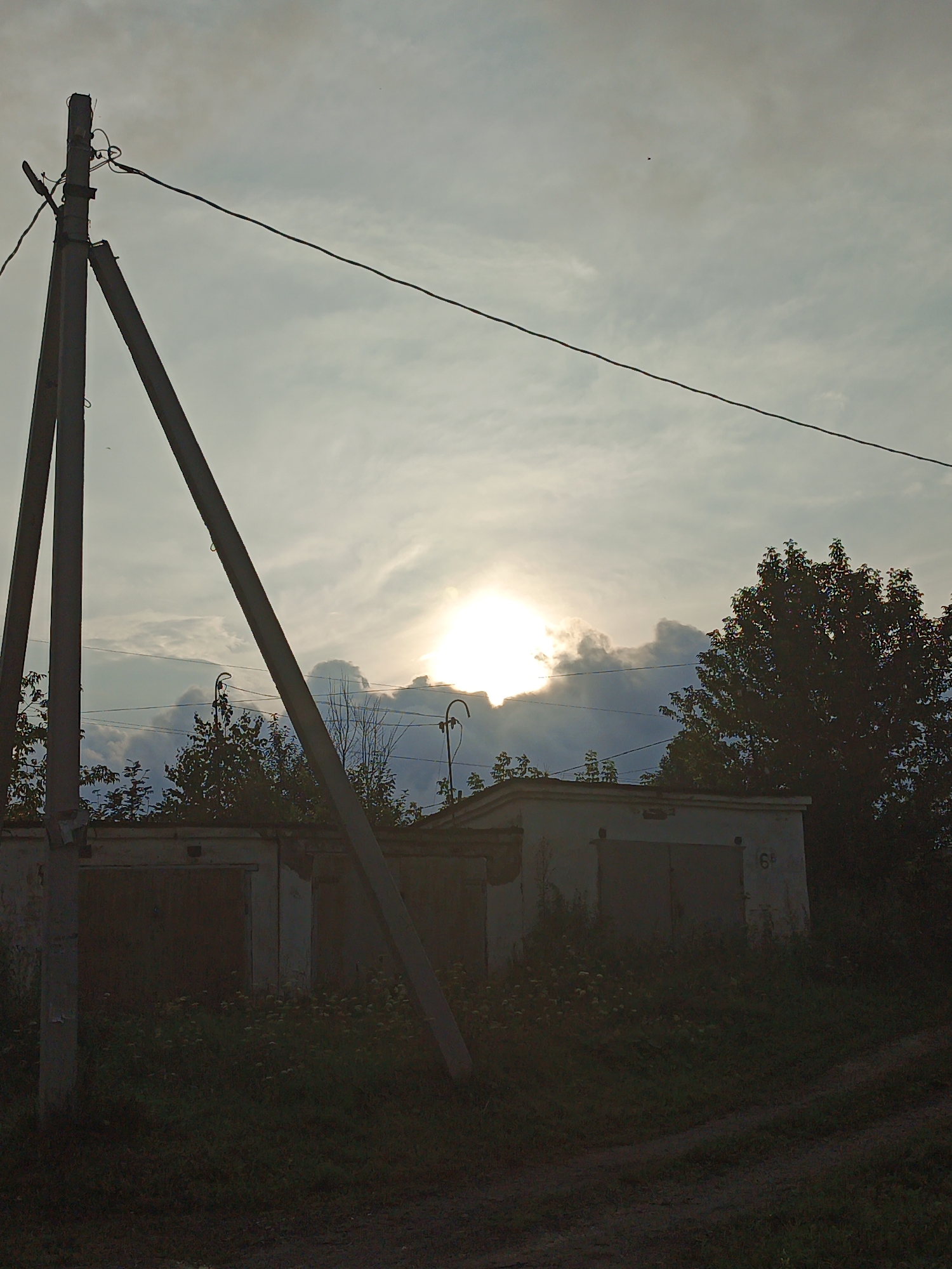 The image depicts a power line pole in the foreground, leaning against a backdrop of a cloudy sky with the sun partially obscured by clouds, creating a bright glow. Behind the pole, a low, white building with garage doors is visible. A number "6" is faintly visible on the side of the building. Trees and vegetation surround the building and extend into the foreground. The overall scene appears to be in a semi-rural or suburban setting, potentially on the outskirts of Medvedyovka, Russia, given the context. The lighting suggests it is either early morning or late afternoon.