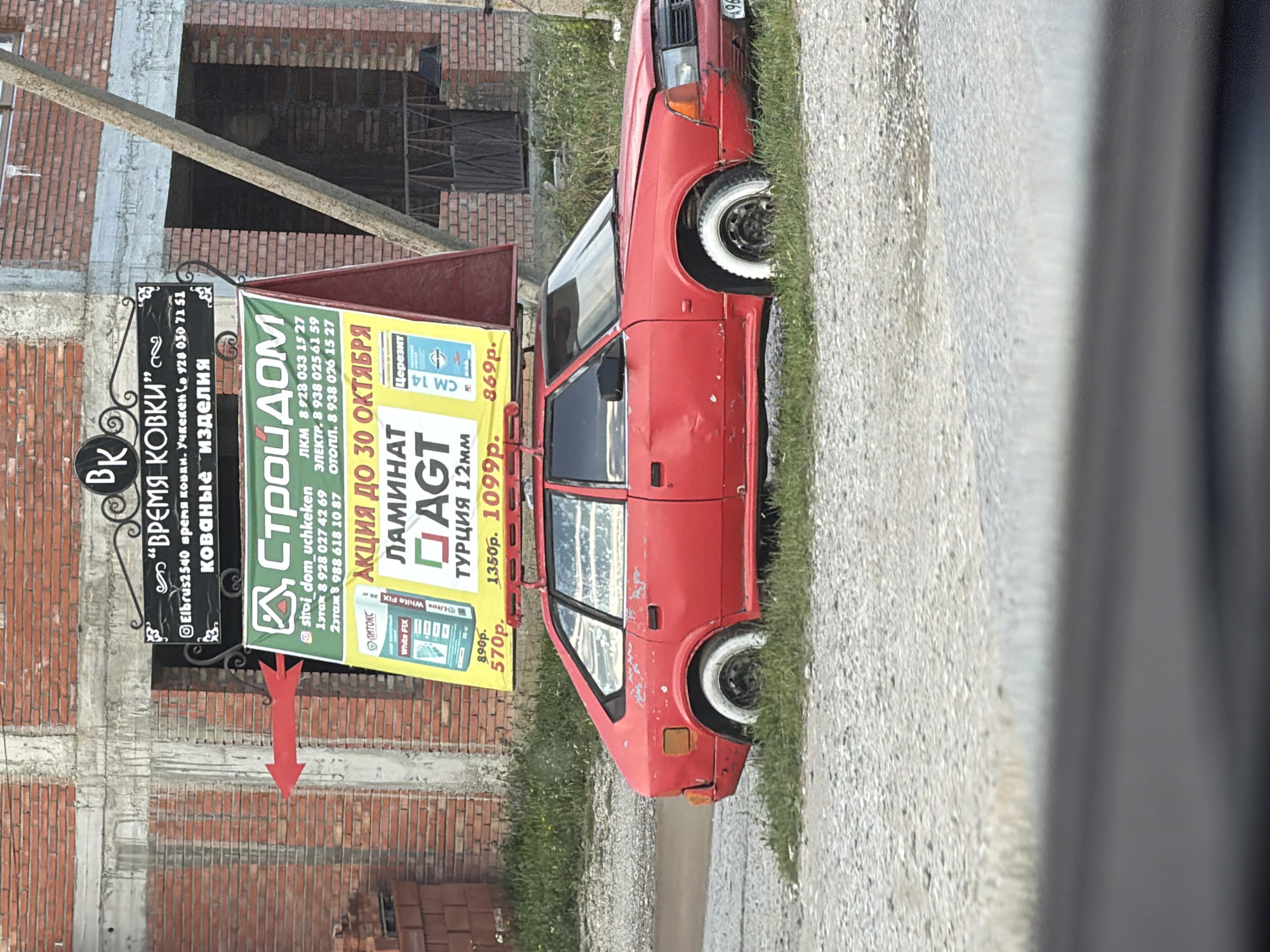 In Джага, Russia, a red car is parked on a gravel surface with grass growing around it. The car has white-painted tires. Behind the car, a large advertising billboard is propped up. The billboard has green and yellow sections, with text and images promoting construction materials. One section advertises "ЛАМИНАТ AGT" (AGT Laminate) and "ТУРЦИЯ 12мм" (Turkey 12mm). Another section shows promotional pricing. To the left of the billboard, a bright red arrow is attached to a brick wall, pointing towards the billboard. Above the billboard, a black sign with gold lettering reads "ВРЕМЯ КОВКИ" (Time of Forging). The background consists of a brick building.