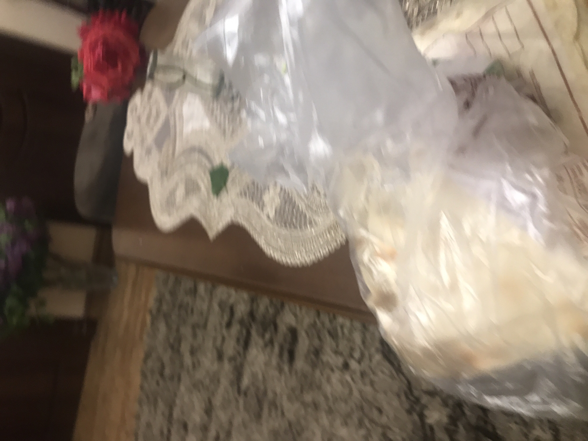 A view from above shows a floral arrangement with red and purple flowers in vases, placed on a dark wood surface. A decorative white lace cloth is draped across part of the surface. A partially transparent plastic bag, containing what appears to be a beige substance, sits next to the floral arrangement. The surface is positioned on a patterned rug. The location in Yerevan, Armenia is not discernible from this image.