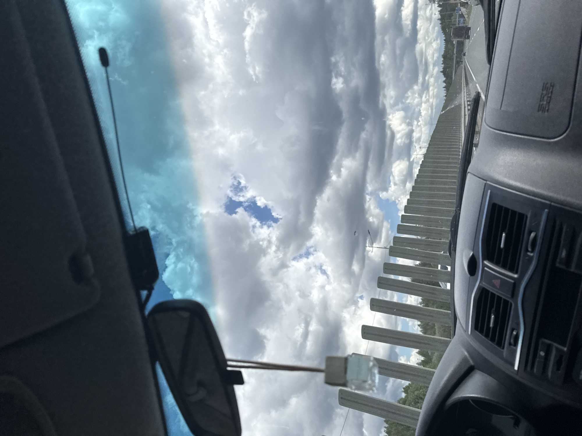 The image shows a view from inside a car, likely on a highway or bridge. The vehicle is moving, as indicated by the blurred scenery. The dashboard and rearview mirror are visible within the car. Outside, the sky is cloudy with patches of blue. A concrete barrier lines the road. Beyond the barrier, trees are visible. Based on the location provided, the image was likely taken in the area near Nagornaya, Russia.