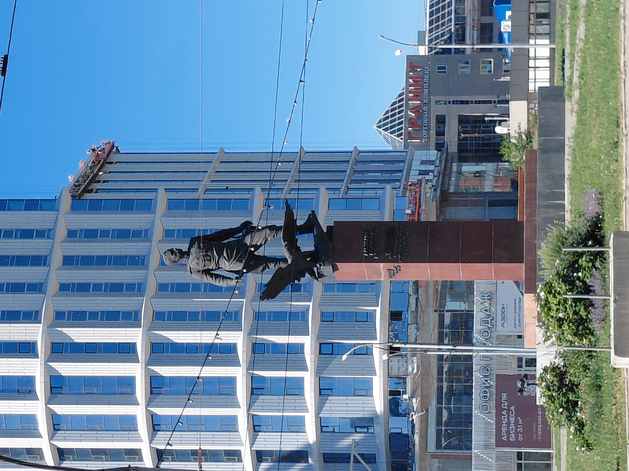 The image depicts a statue of a man in military uniform standing on a pedestal, with an eagle at his feet. Behind the statue, a modern, curved building with many windows dominates the background. The sky is clear and blue. In the middle ground, there is a sign with Russian text that reads "ГРАНИТ" (Granit) and "ТОРГОВЫЙ КОМПЛЕКС" (Shopping Complex). There are also signs for "ОФИС ПРОДАЖ" (Sales Office) and "АРЕНДА ДЛЯ БИЗНЕСА" (Rent for Business). The scene appears to be in a city. The statue is likely a monument commemorating a historical figure or event. The modern architecture of the building suggests a contemporary urban setting. The presence of commercial signage indicates an active area with businesses. The overall impression is one of a public space in a city, possibly a plaza or square, where historical and modern elements coexist. The statue is located in front of the building of the Novosibirsk State University of Architecture and Civil Engineering, with the statue of Ivan Ivanovich Lavrentiev.