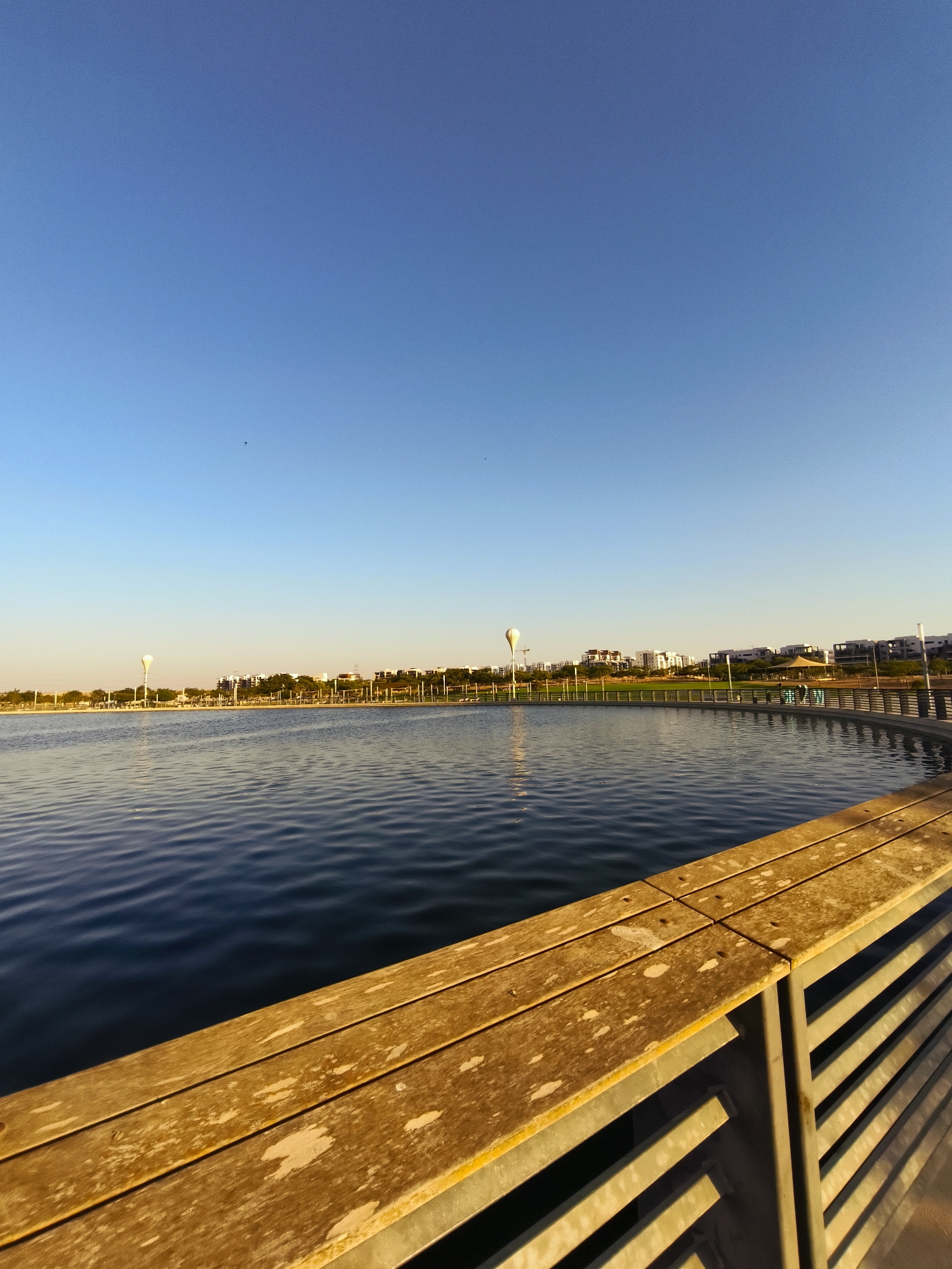 The image captures a serene view of the Beer Sheva River Park in Beer Sheva, Israel. The foreground is dominated by a weathered wooden railing and seating area, casting long shadows on the slatted metal structure beneath. Beyond this, a calm body of water reflects the clear, bright blue sky. In the middle ground, a curved pathway lined with a fence runs along the edge of the water. Across the river, a vibrant cityscape unfolds with modern buildings, lush green spaces, and trees. A distinctive, large white balloon-shaped sculpture stands tall on the left side, illuminated by the warm glow of the setting or rising sun. The overall scene conveys a peaceful atmosphere, characteristic of a park at the golden hour.