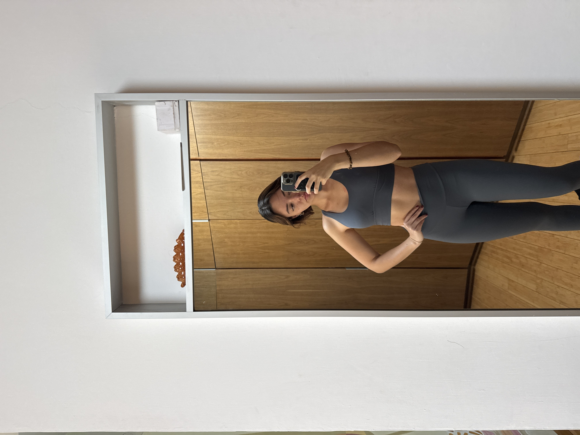 A woman stands facing a mirror, holding a smartphone to take a picture of herself. She is wearing a gray sports bra and leggings. The mirror is attached to a wall with light-colored paneling. To the left of the mirror is a white wall with a decorative item mounted on it. A wooden floor is visible in the reflection. The location is in San Bernardino Tlaxcalancingo, Mexico.
