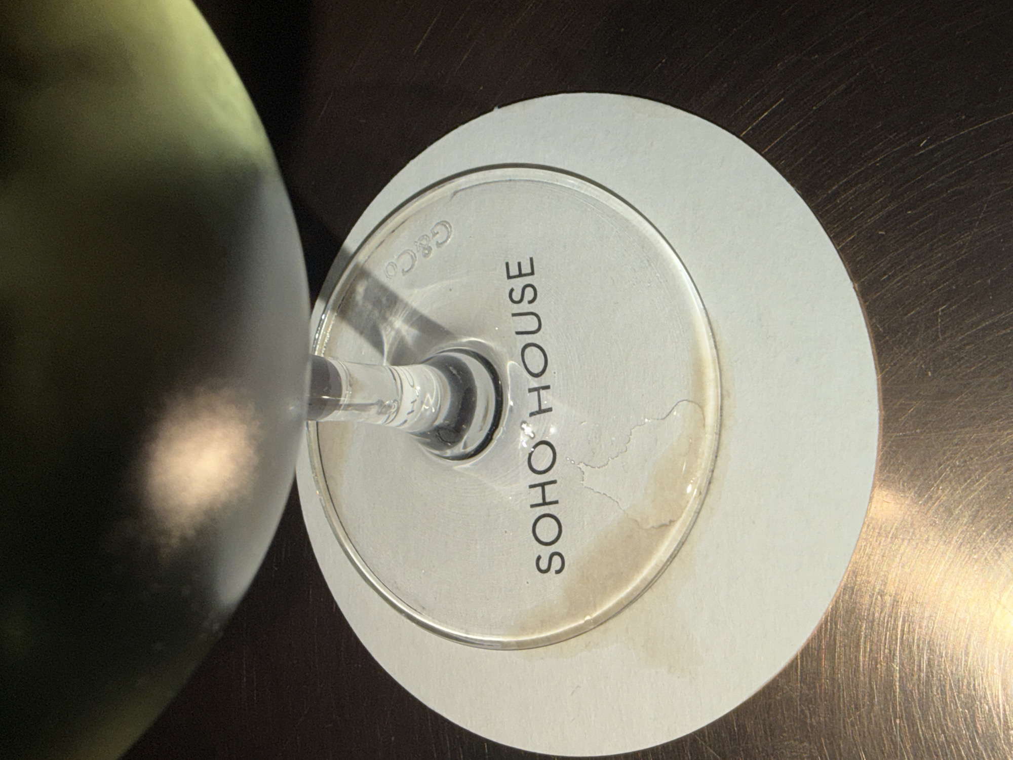 The image shows a wine glass sitting on a white, circular coaster on a reflective, possibly metal, surface. The base of the glass is imprinted with the words "SOHO HOUSE" in black lettering, as well as "G&Co" near the edge of the glass. There are some liquid stains on the coaster and base of the glass. Part of a green wine bottle is visible in the left side of the frame. Soho House is a members club with multiple locations, including in London, United Kingdom.