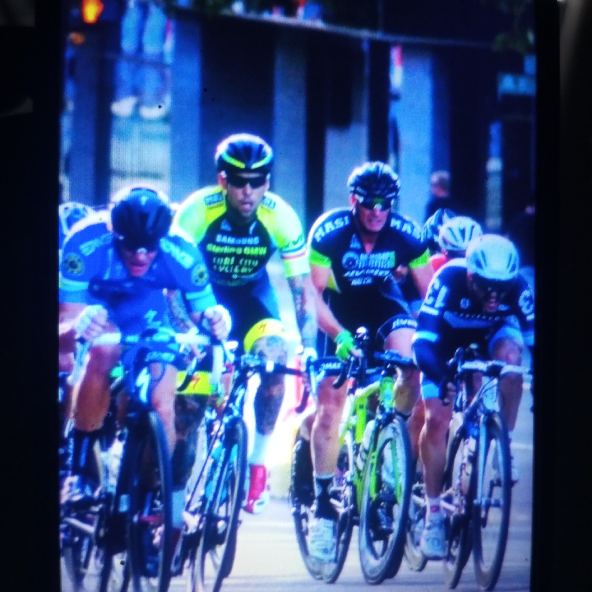 The image captures a dynamic, close-up view of a professional cycling race. Three male cyclists are prominently featured, all wearing helmets and cycling gear. The cyclist in the foreground on the left is wearing a blue and white jersey with the number 1 on his bike. The cyclist next to him, slightly behind and to the right, wears a neon yellow and black jersey and dark sunglasses. To the right of the frame, another cyclist in a black and green jersey is visible, also wearing a helmet and dark glasses. Their bikes are equipped with aerodynamic wheels and visible sponsorships. The cyclists appear to be in a competitive stride, leaning forward over their handlebars. The background is blurred, suggesting motion, but hints of urban architecture and other blurred figures indicate a public setting within a city. The overall lighting suggests it is daytime, possibly overcast or in a shaded area. There is no information in the image that suggests the location is Cemani, Indonesia.