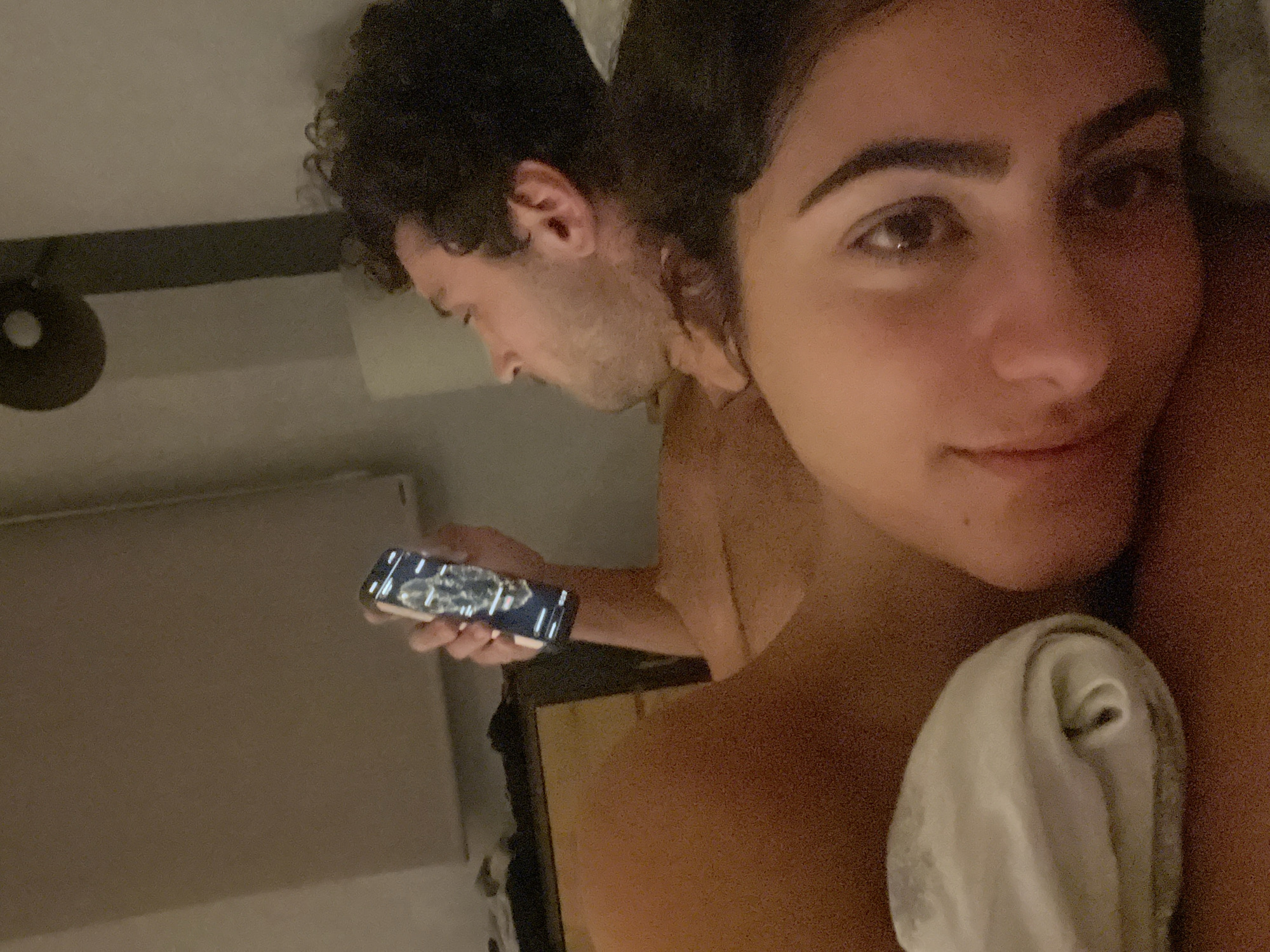 Two individuals are lying in bed. The person in the foreground is a woman, whose face is partially visible, looking directly at the camera with her eyes open. She has dark eyebrows and is lying on her side. Behind her, to the right, is a man, also lying on his side and looking upwards and to the left. He has curly dark hair and a beard. The man is holding a smartphone in his right hand, which is displaying a screen with an image. The environment appears to be a bedroom, with a wall and a light fixture visible in the background. The lighting is dim. There are no indications or clues that would suggest the location is Madrid, Spain.