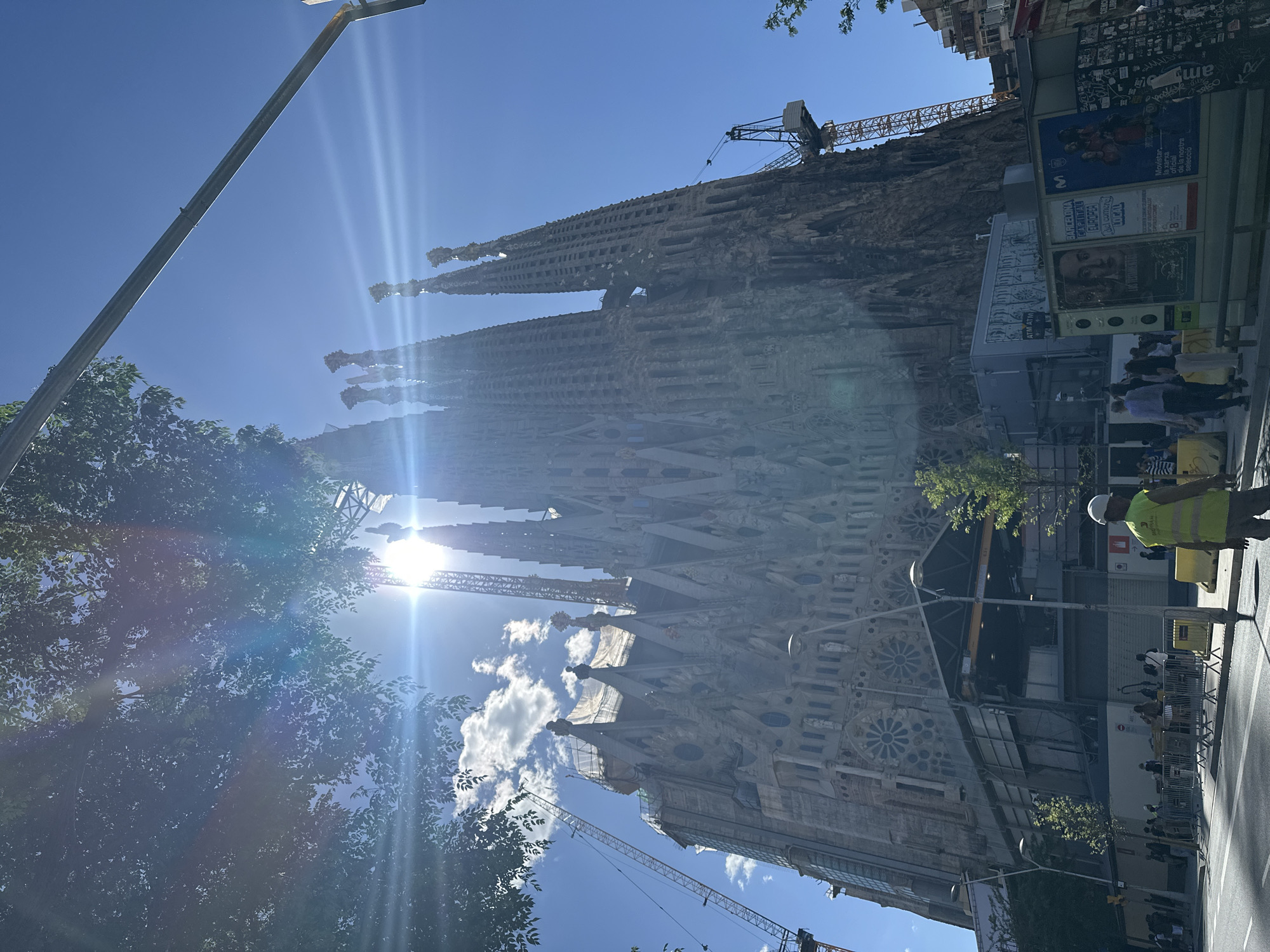 This is an eye-level outdoor shot of the Sagrada Familia in Barcelona, Spain. The tall, ornate spires of the basilica rise high into a bright, clear blue sky. Construction cranes are visible, indicating ongoing building work. In the foreground on the right is a street with pedestrians, and a construction worker wearing a helmet and a yellow vest. A bus stop or advertising panel is nearby. The basilica is partially obscured by the sun and some trees on the left.