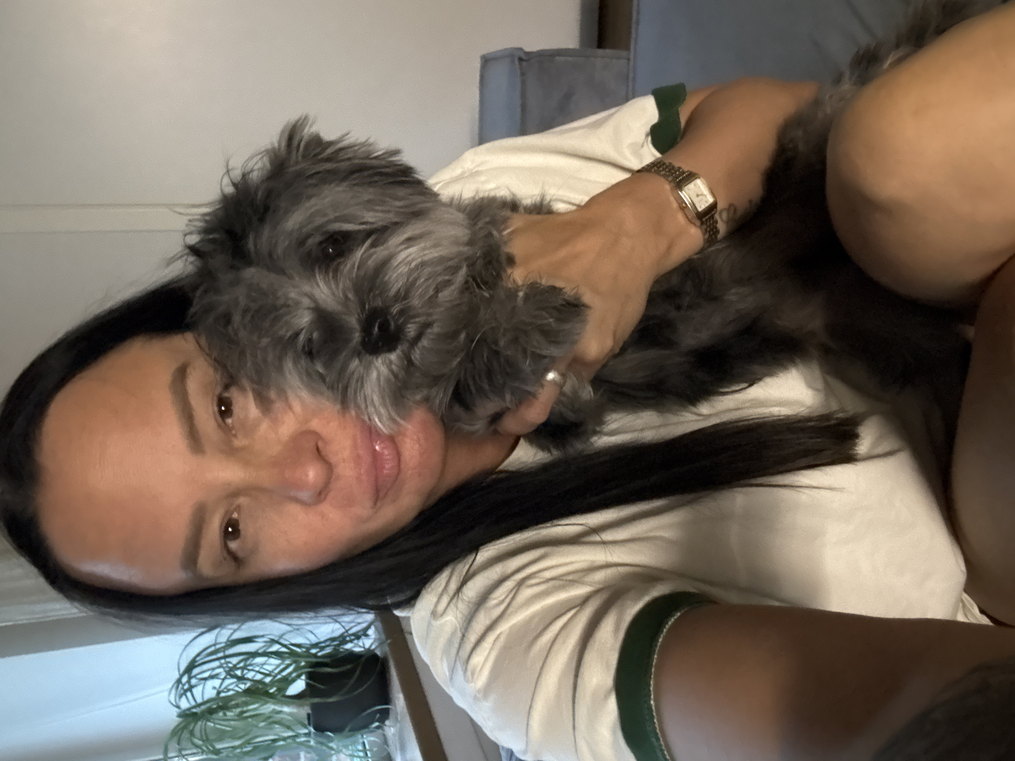 A woman is holding a gray, shaggy dog close to her face in an indoor setting. The woman has long, dark hair, light skin, and is wearing a white t-shirt with green trim on the sleeves. She is wearing a gold watch on her left wrist. The dog is mostly gray with darker features around its face. The background is blurred, but includes a gray sofa or cushion and what appears to be a plant in a pot. The photo appears to be a selfie. There is no information in the image to suggest this photo was taken in Chaikovsky, Russia.