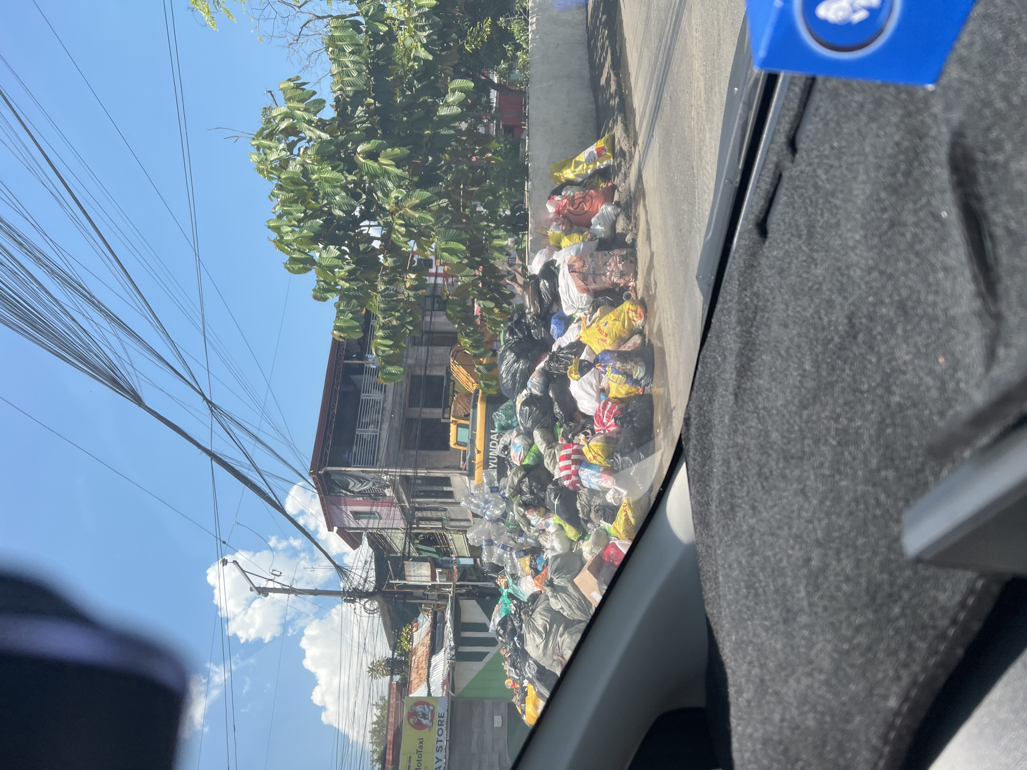 The image presents a street view in Davao City, Philippines, captured from inside a vehicle on a bright, sunny day. A substantial accumulation of garbage occupies the lower-mid section of the frame, forming a large, irregular pile along the roadside. This heap comprises numerous plastic bags of various colors including black, white, yellow, red, and orange, many appearing full. Interspersed within the pile are clear plastic bottles, cardboard boxes, and other discarded materials.
In the background, multiple buildings line the street. To the left, a single-story structure with a corrugated metal roof displays partial signage reading "MotoTaxi" and "AY STORE." Adjacent to it and further right, a two-story building with a red roof and exposed concrete construction is visible. Partially obscured by the garbage pile and this building is a yellow Hyundai excavator. To the right of the buildings, a large tree with dense green foliage stands, extending above a light-colored concrete retaining wall that runs along the roadside.
The sky is clear blue with several scattered white cumulus clouds. A complex network of overhead utility lines and cables spans across the upper portion of the scene, connected to wooden utility poles. The road surface in the foreground and midground is asphalt. The immediate foreground shows the dark grey, felt-covered dashboard or parcel shelf of the vehicle from which the photograph was taken. A blue box with a white circular logo is partially visible on the far right of the dashboard. No individuals are visible in the scene.