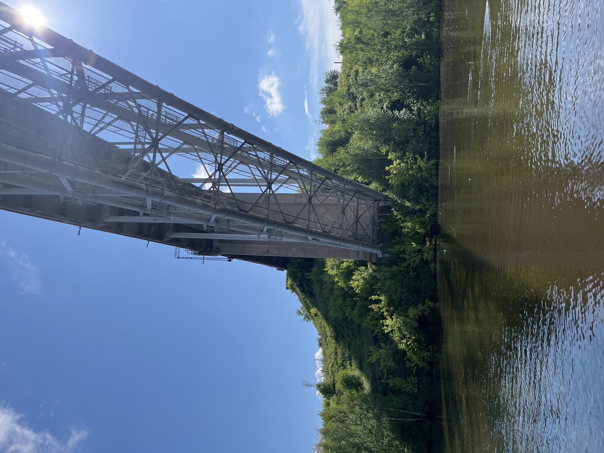 The image shows a view from a body of water, likely a river, looking up at a large, elevated bridge with a complex metal structure. The bridge arches over a green, tree-covered hill. The sky is blue with a few scattered white clouds. Sunlight is visible in the upper right corner, casting lens flare onto the image. The water reflects the sky and the bridge, with ripples on its surface. In the background, on the top of the hill, there appears to be some infrastructure, possibly telecommunication towers. The vegetation is lush and green, indicating a temperate season. There are no people or animals visible in the image. The scene suggests an outdoor, natural setting enhanced by man-made infrastructure, characteristic of a city with both urban development and natural landscapes, such as Kamensk-Uralsky, Russia, known for its industrial heritage and surrounding nature.