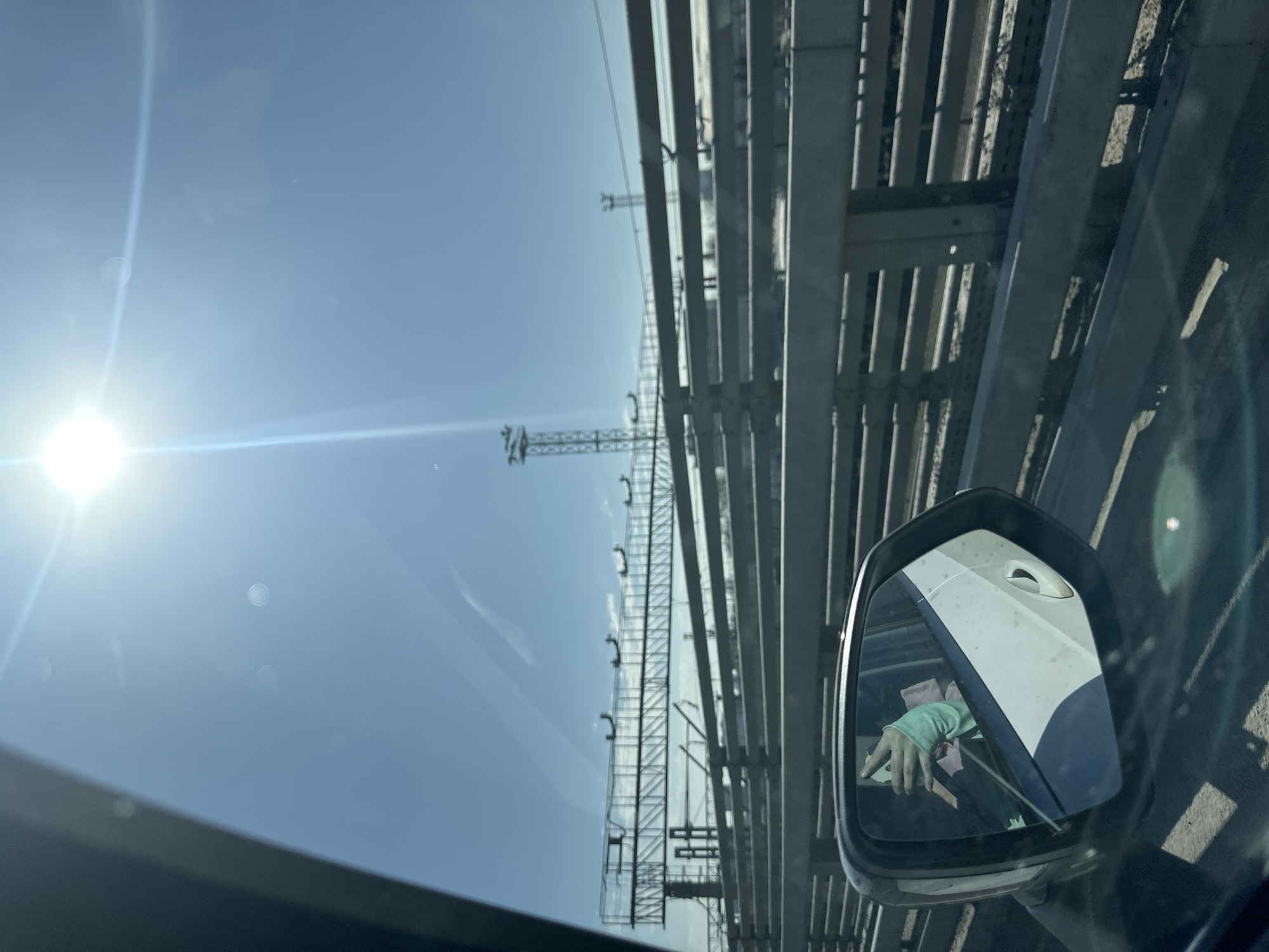 The image, taken from inside a car, shows an exterior view through the car window and a reflection in the side mirror. The sky is bright blue with a visible sun. An elevated structure, possibly a bridge or industrial framework, dominates the view through the window, consisting of gray metallic beams. A telecommunications tower is also visible. The side mirror reflects the interior of the car, with a person's hand visible wearing a light green sleeve. The location and details of Moscow, Russia are not discernable from the image.