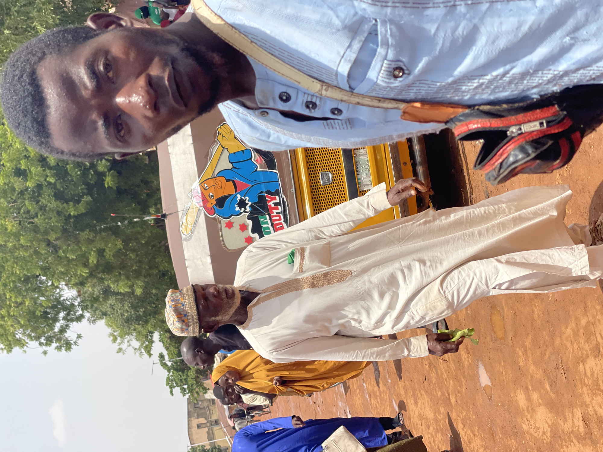 The image captures a street scene in Zaria, Nigeria. A man in a light blue, button-down shirt and with a black bag is in the foreground, partially out of focus. Behind him, another man in a white robe with a patterned cap holds green leaves. In the background, a yellow truck has a cartoon drawing. Several other people are visible in the background wearing traditional African clothing in shades of blue, yellow, and black. Trees are in the left background. The ground is reddish-brown.