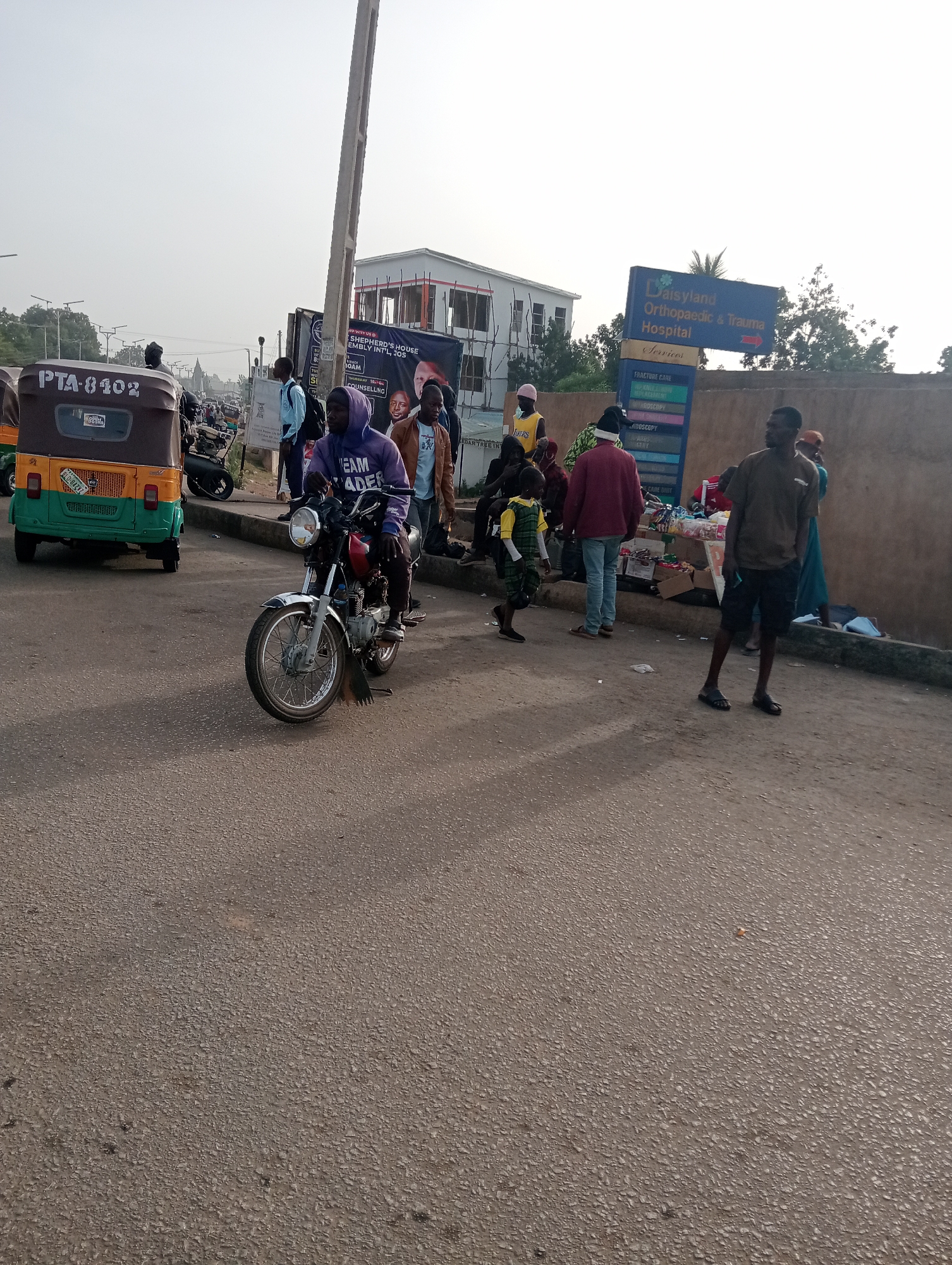 The image depicts a street scene in Jos, Nigeria, adjacent to the "Daisyland Orthopaedic & Trauma Hospital." A motorized tricycle with license plate "PTA-8102" is parked on the left side of the frame. A man on a motorcycle is positioned in the center of the road, facing the camera. A group of people are gathered near the hospital sign and along the sidewalk, some appearing to be engaged in commerce with a small street vendor selling goods. A banner with the text "SHEPHERD'S HOUSE ASSEMBLY INT'L, JOS" is partially visible. The overall environment suggests a bustling, urban setting.