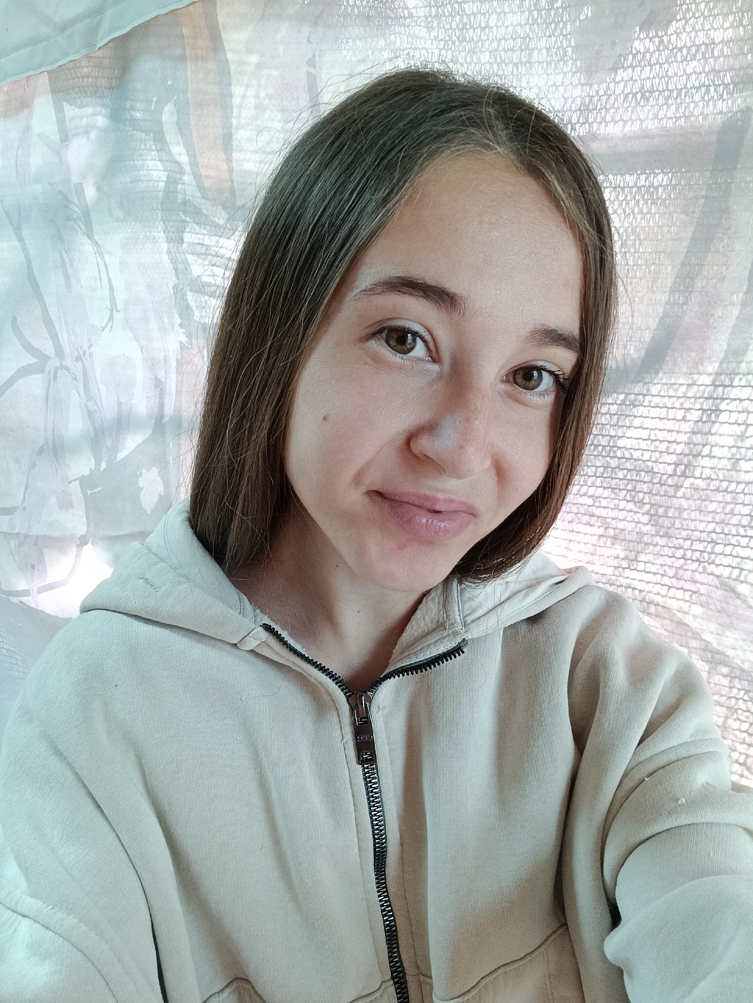 A young woman with long, dark brown hair and brown eyes is captured in a close-up, selfie-style photograph. She is wearing a light beige zip-up hoodie. The background consists of a light-colored, textured fabric, possibly a curtain or a wall covering, with a floral or abstract pattern visible in soft pastel colors. The lighting suggests an indoor setting, potentially near a window given the diffused light and the texture of the background material. The image is taken from a slightly elevated angle, looking down at the subject. There are no identifiable landmarks or objects that would indicate the specific location within Vypasne, Ukraine.