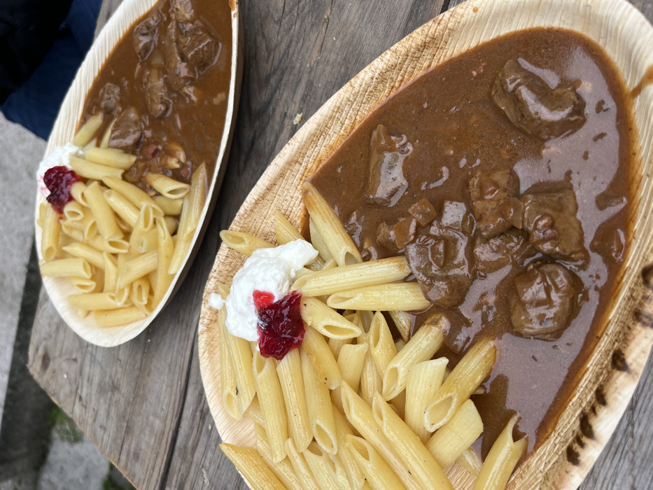 This image depicts two meals served on disposable, natural-fiber, boat-shaped plates, positioned on a weathered wooden surface. Each plate contains an identical serving: a portion of light-yellow penne pasta occupying approximately half the plate, alongside a generous portion of a dark brown meat stew containing cubed meat pieces and what appear to be diced vegetables. A dollop of white sour cream or similar dairy product is placed on the pasta, topped with a small amount of reddish-purple fruit preserve or jam. The plates are arranged diagonally, with one in the foreground and the other slightly behind and to the right, partially obscured. The wooden surface exhibits grain patterns and minor wear. In the upper right background, a dark blue fabric, likely clothing, is partially visible, suggesting the presence of an individual or their belongings beyond the immediate dining surface. The background is softly blurred, indicating an outdoor setting in Obermoschel, Germany.