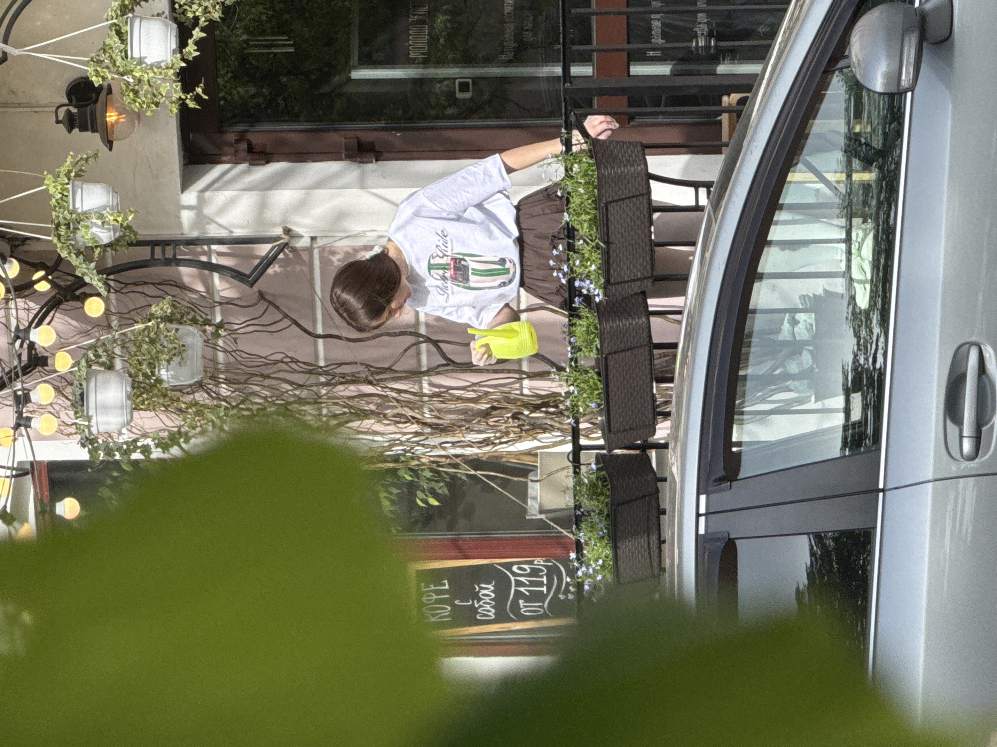 A woman is watering flowers in a planter near a building with a sign in Russian that translates to "Coffee to go from 119r". The woman is wearing a white t-shirt with a race car design and a brown skirt. She is holding a yellow watering can. The location is in the city of Kazan, Russia. A silver van is parked on the street. There are string lights and potted plants. The lower portion of the image is obscured by green leaves.