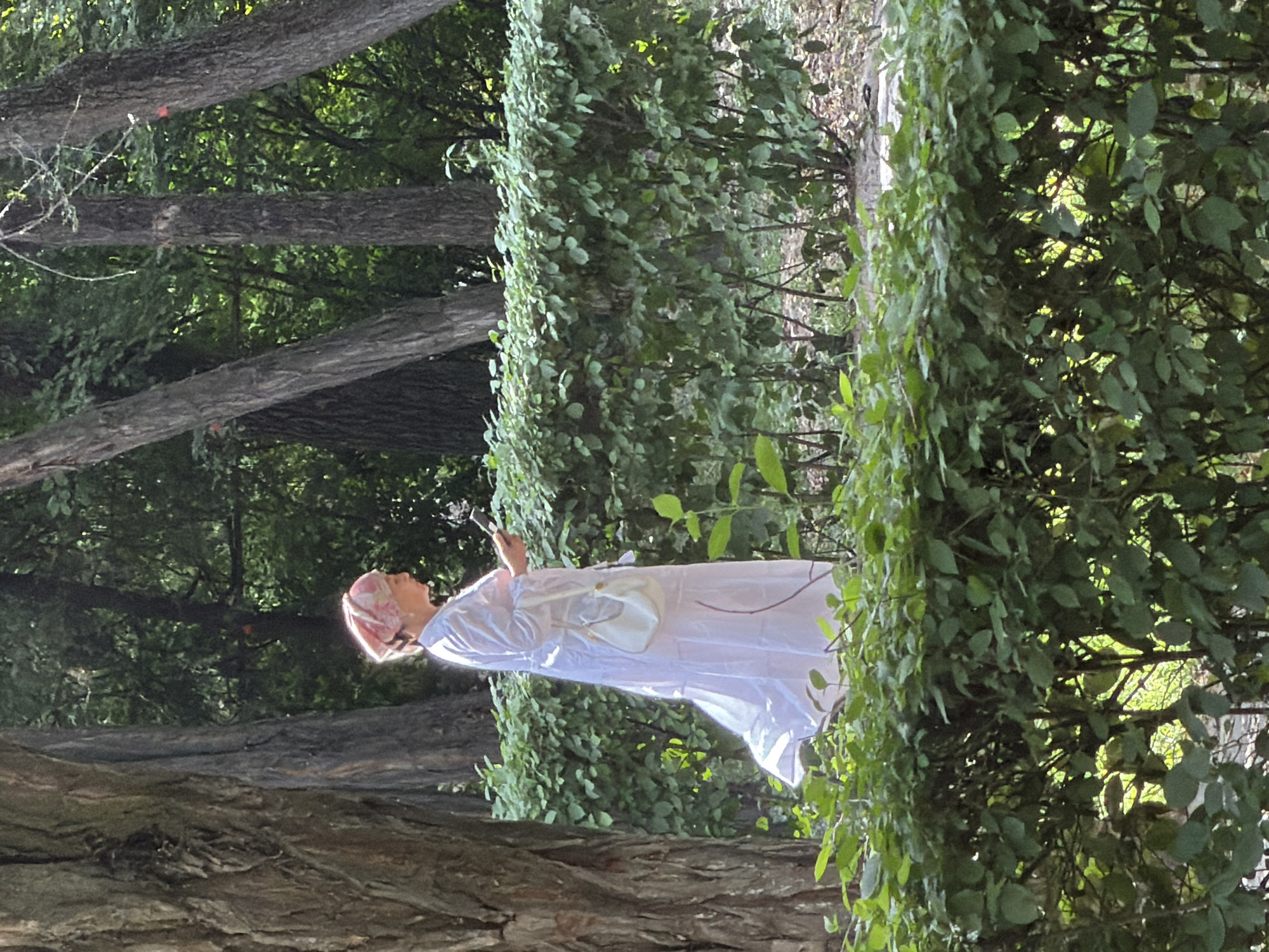 A woman in a white dress and pink headscarf stands in a park, looking at her phone. She is holding a small white purse. The woman is surrounded by lush green foliage, including trees and bushes. Sunlight filters through the leaves, creating a dappled effect on the ground. The scene appears to be in a public park, possibly in a city given the maintained hedges.