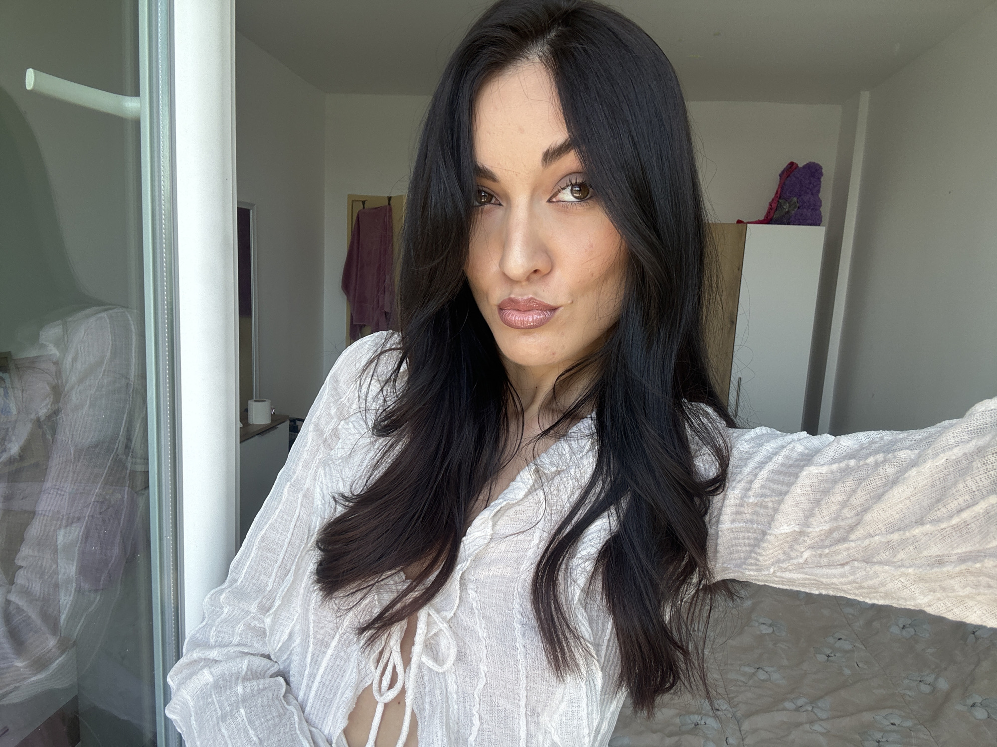 A young woman with long, dark, wavy hair is taking a selfie indoors. She is wearing a white, sheer, textured blouse with a tie-front closure. Her makeup is visible, including lipstick. The background shows a window with a white frame and a view of what appears to be a domestic interior. A white dresser or cabinet is visible to the right, with a purple object on top. The lighting suggests it is daytime. The image is not taken in Burgas, Bulgaria.