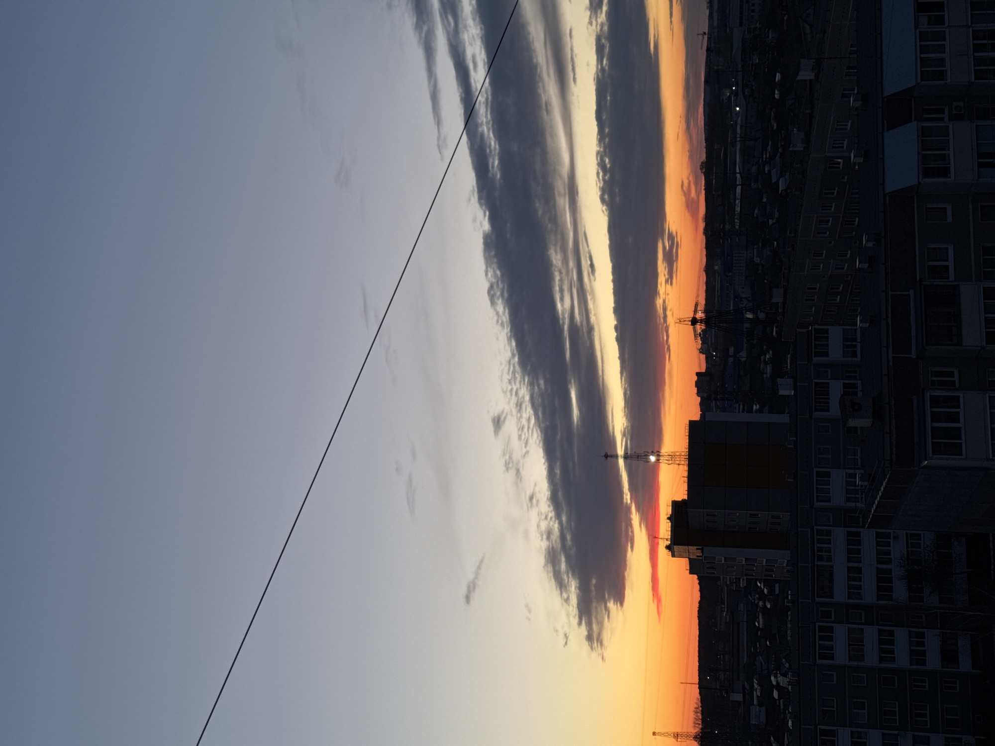The image captures a sunset in what appears to be the city of Chaikovsky, Russia. Tall apartment buildings are silhouetted against the evening sky. The sky is a mix of blue, white, orange, and red hues, indicating the setting sun. Clouds stretch across the sky, some reflecting the colors of the sunset. There's a thin cable crossing diagonally from the top left. Construction cranes are visible on the horizon. The urban environment is evident through the presence of buildings and infrastructure. The image does not contain people.