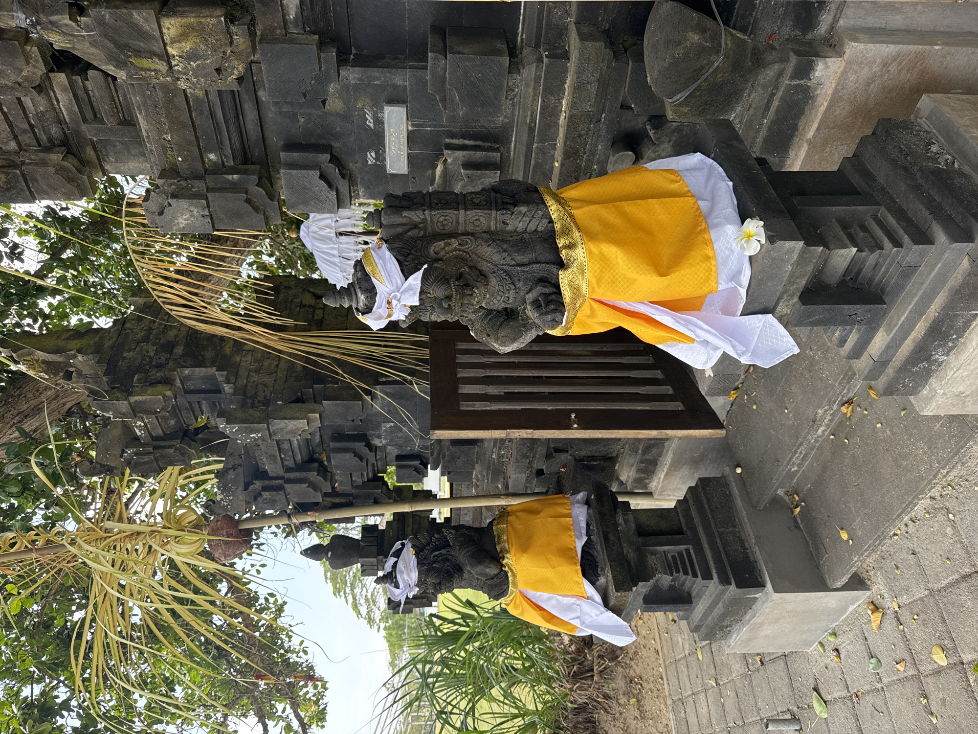 Two stone statues, adorned with yellow and white cloths, stand guard at the entrance of a dark stone structure, possibly a temple or shrine, in Kuta Selatan, Indonesia. The statues are weathered and detailed, depicting figures with fierce expressions. A white flower rests on the base of the statue on the right. The entrance is a wooden gate with vertical slats. Palm fronds hang overhead, suggesting a tropical environment. The ground is paved with square stone tiles, scattered with fallen leaves. In the background, lush green grass and trees are visible. The overall scene conveys a sense of cultural heritage and spiritual significance within a tropical setting.